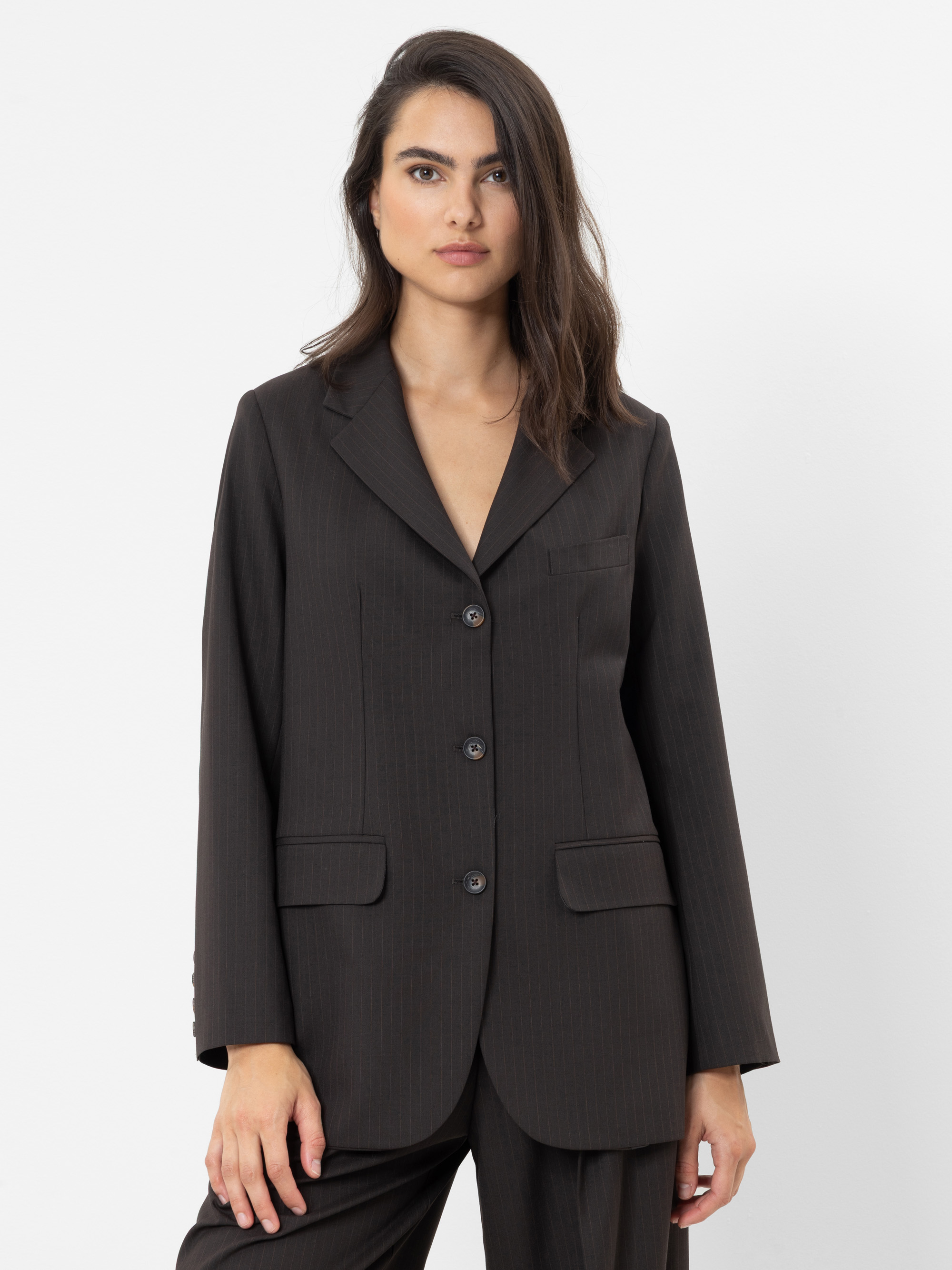 Eine Person mit langen, dunklen Haaren, die ein dunkles, Nadelstreifen-Anzugjacke und eine dazu passende Hose trägt und vor einem schlichten weißen Hintergrund steht.