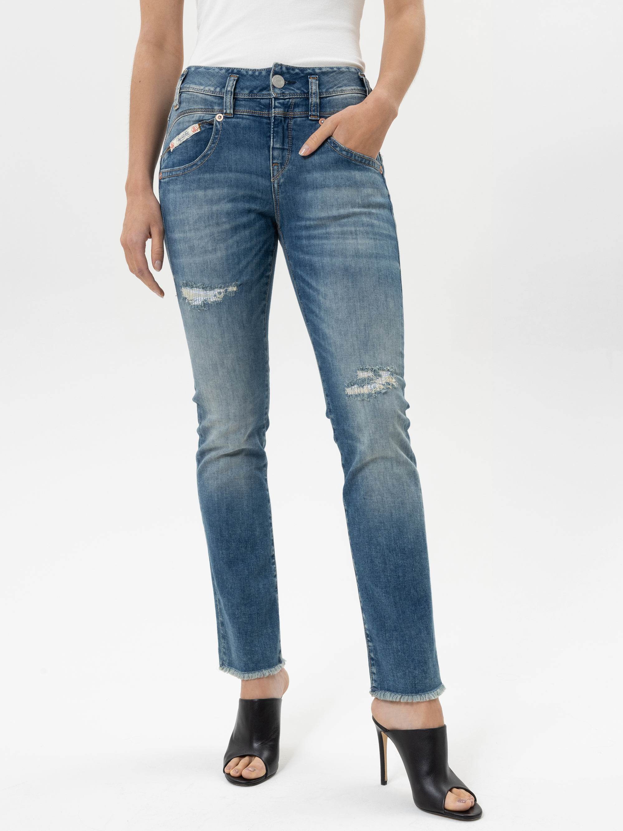 Eine Person trägt eine abgenutzte Bluejeans mit ausgefransten Säumen und ein weißes Oberteil und steht mit einer Hand in einer Tasche. Sie trägt außerdem schwarze, hochhackige, offene Schuhe vor einem schlichten weißen Hintergrund.