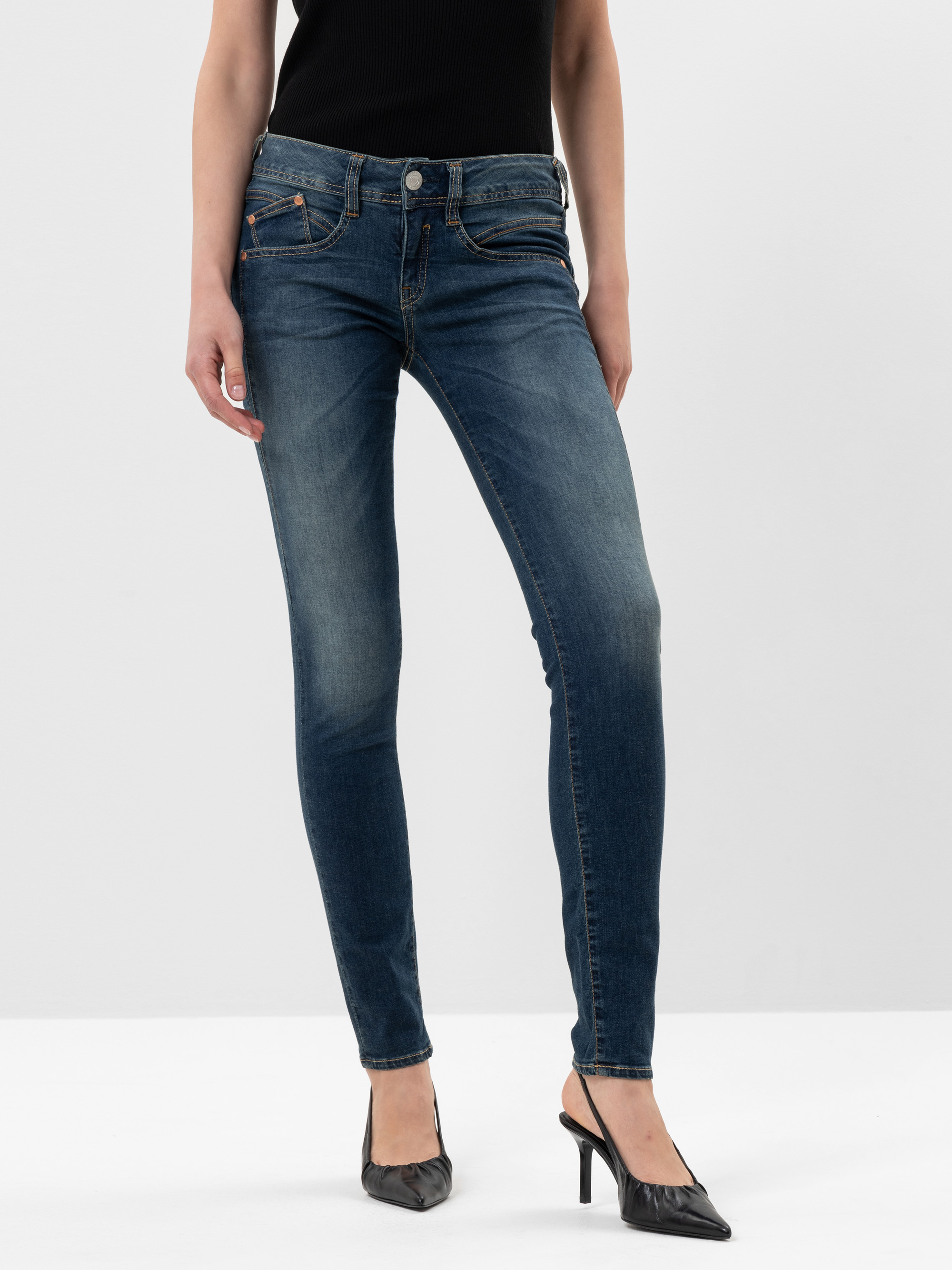 Eine Person, die ein schwarzes ärmelloses Oberteil, blaue Röhrenjeans und schwarze spitze Absätze trägt, steht vor einem schlichten weißen Hintergrund. Das Foto zeigt den Körper von den Schultern bis zu den Füßen.