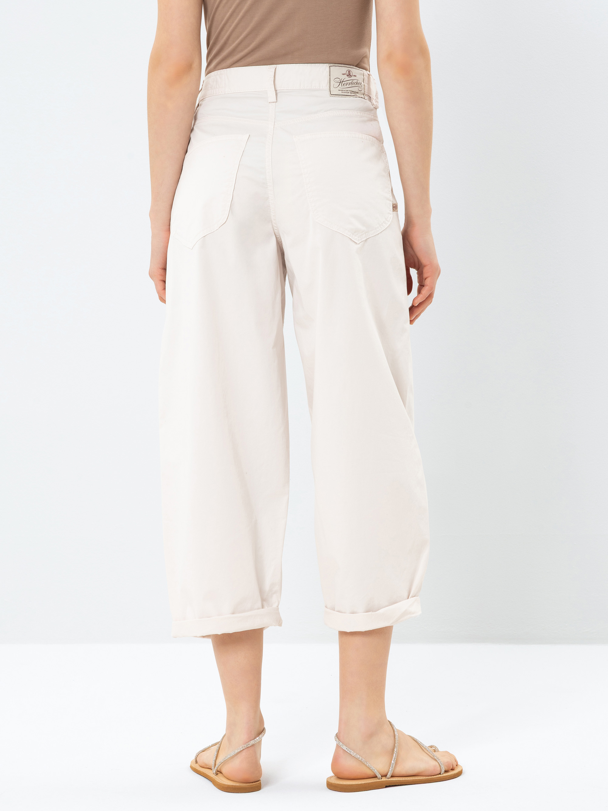 Eine Person steht mit dem Gesicht zur Wand und trägt eine hellbeige Hose mit weitem Bein und Bündchen am Saum sowie ein hellbraunes ärmelloses Oberteil, dazu flache Sandalen. Der Hintergrund ist einfarbig und weiß.