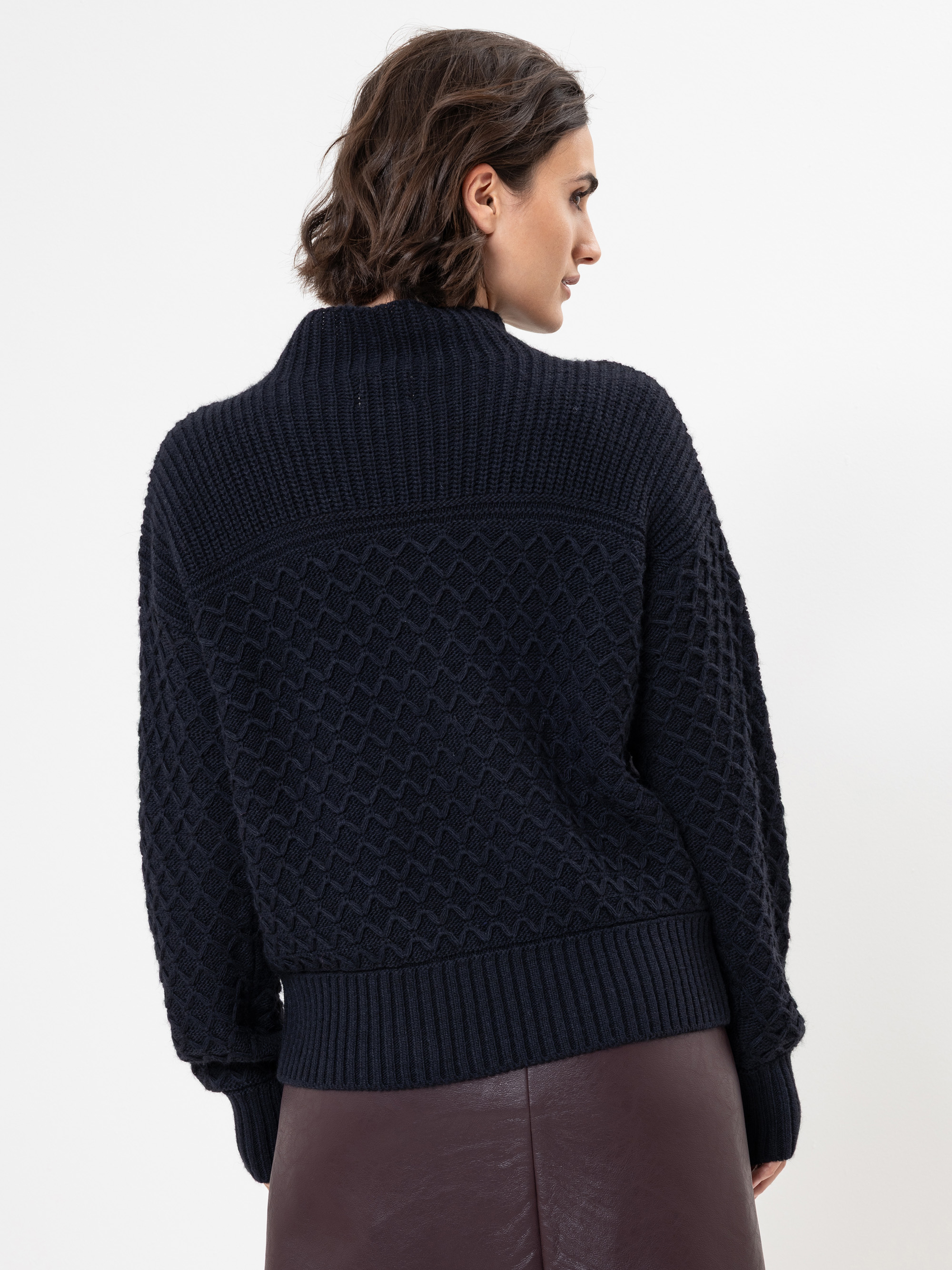 Eine Frau mit kurzen braunen Haaren, von hinten gesehen, trägt einen dunkelblauen strukturierten Strickpullover und einen hochtaillierten dunkelbraunen Rock vor einem schlichten weißen Hintergrund.