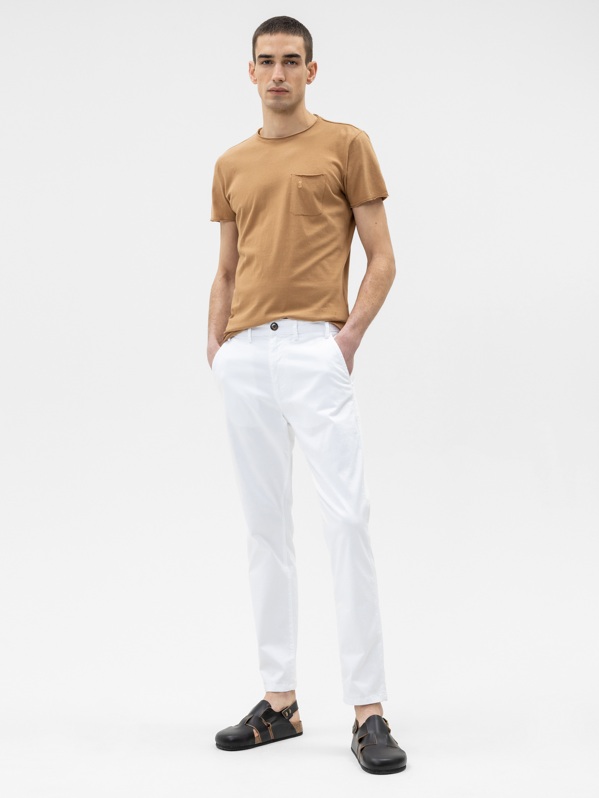 Ein Mann steht vor einem schlichten weißen Hintergrund, trägt ein braunes kurzärmeliges T-Shirt, eine weiße Hose und schwarze Sandalen, hat die Hände in den Taschen und einen neutralen Gesichtsausdruck.