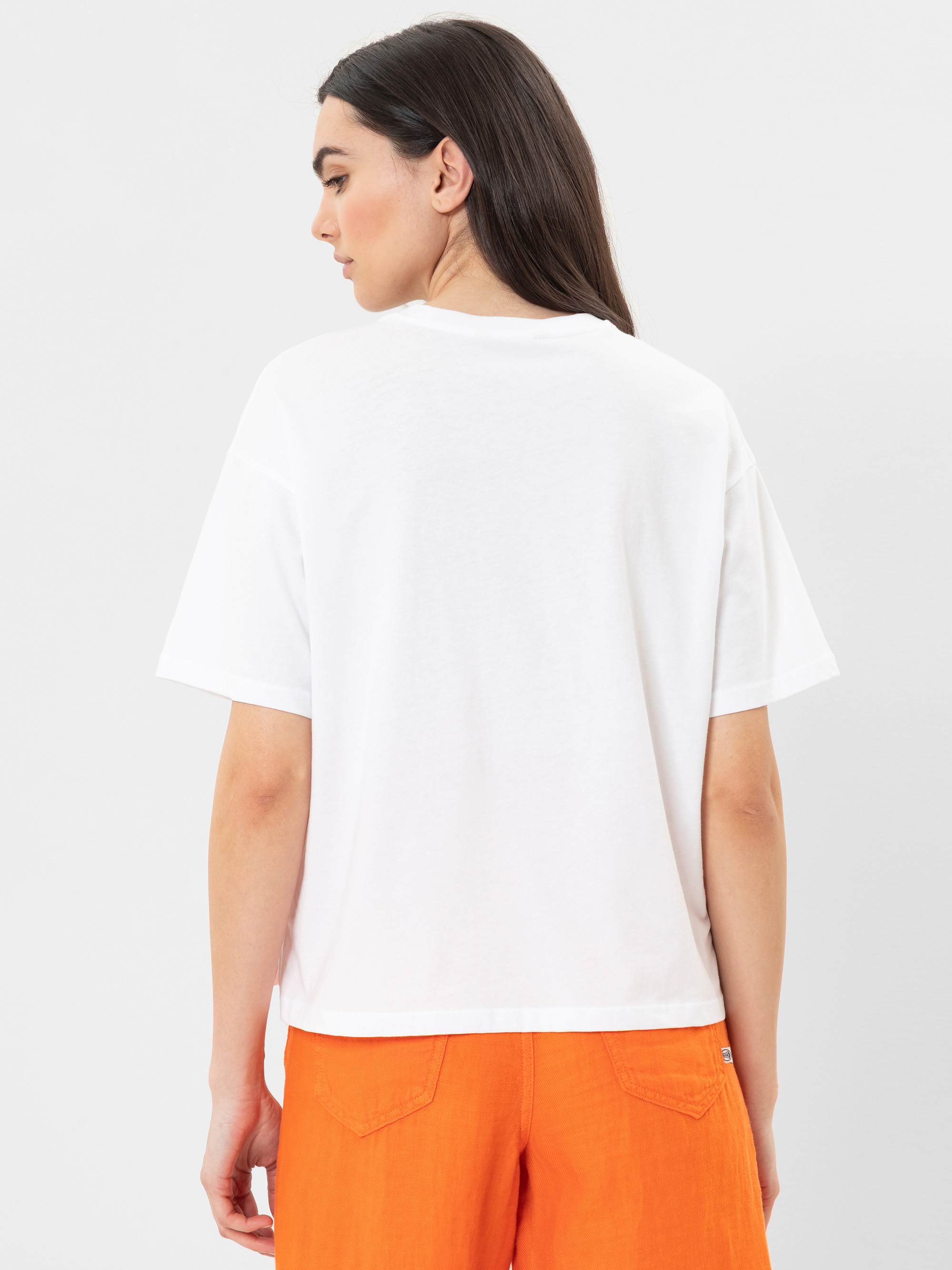 Eine Frau mit langen dunklen Haaren steht mit dem Gesicht zur Kamera, trägt ein schlichtes weißes kurzärmeliges T-Shirt und leuchtend orangefarbene Shorts vor einem hellen Hintergrund.