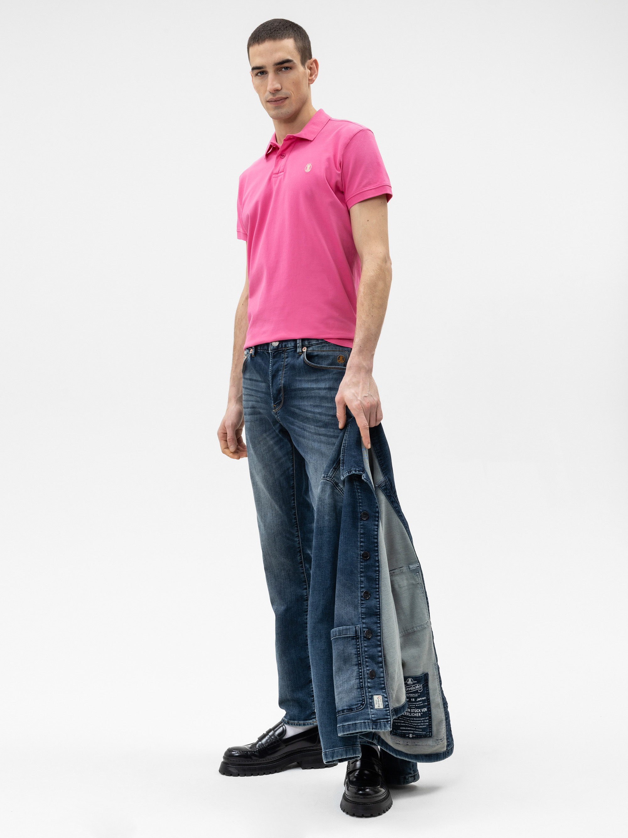 Ein Mann in einem hellrosa Poloshirt, blauen Jeans und schwarzen Schuhen steht mit einer Jeansjacke in der Hand vor einem schlichten weißen Hintergrund.