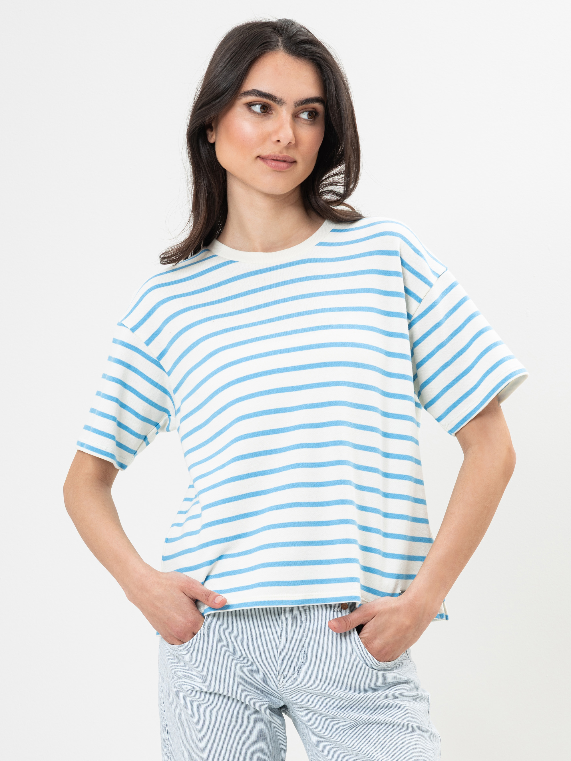 Eine Frau mit langen dunklen Haaren trägt ein weißes T-Shirt mit hellblauen Querstreifen und eine helle Jeanshose und steht mit den Händen in den Taschen vor einem schlichten weißen Hintergrund.