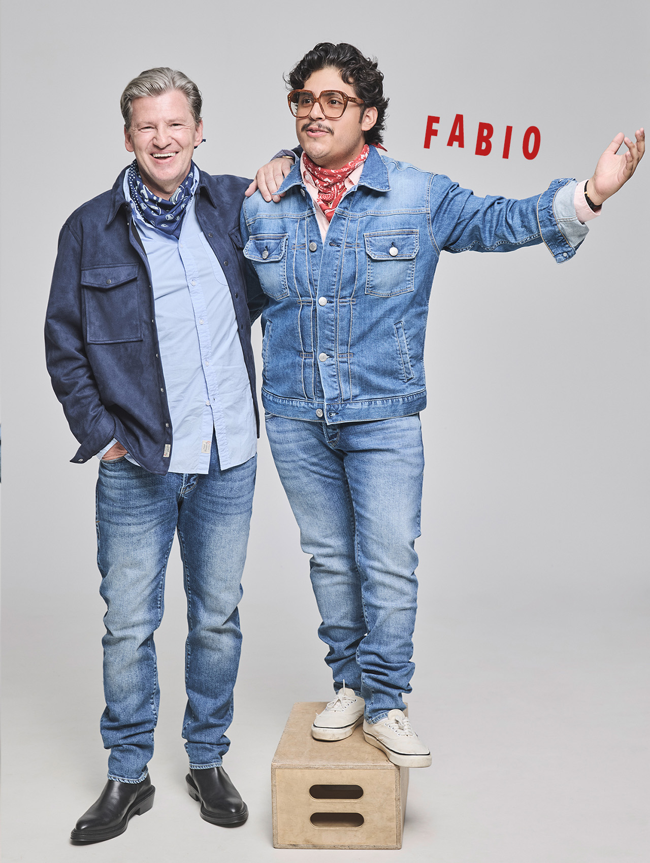 Zwei Männer posieren zusammen in Freizeitkleidung und Jeansjacken. Einer steht mit ausgestrecktem Arm auf einer Holzkiste, über ihm steht in roter Schrift FABIO. Der andere steht neben ihm, beide lächeln in die Kamera vor einem schlichten Hintergrund.