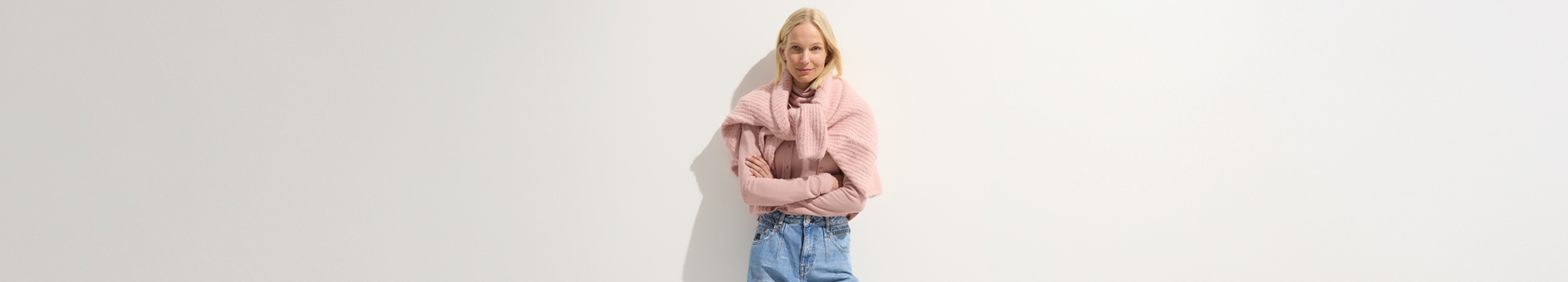 Eine Person steht vor einem schlichten weißen Hintergrund, lächelt leicht, hat einen rosa Pullover über die Schultern gelegt und die Arme verschränkt. Sie trägt ein hellrosa Oberteil und blaue Jeans.