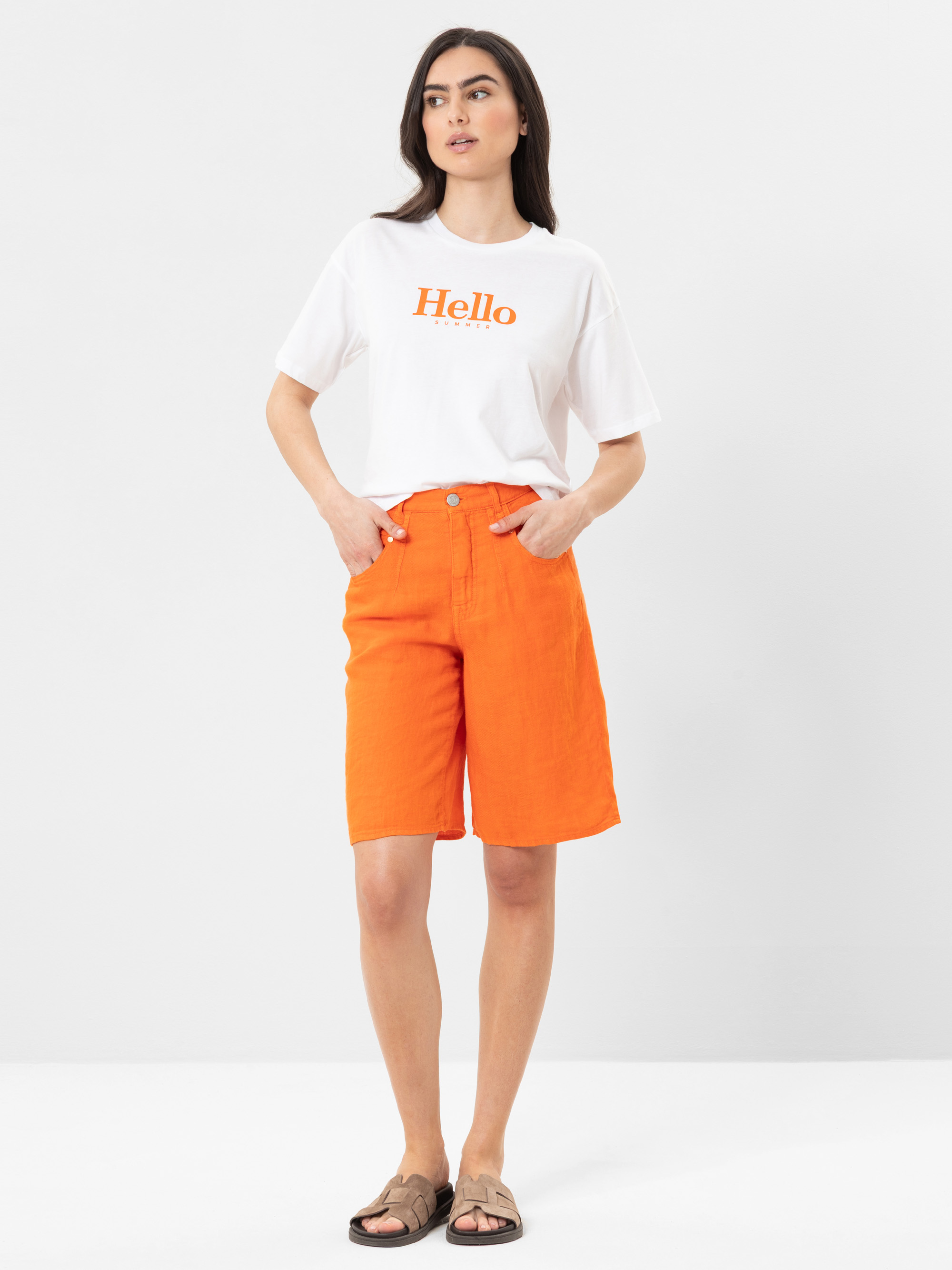 Eine Frau steht vor einem schlichten Hintergrund und trägt ein weißes Hello-T-Shirt, leuchtend orangefarbene knielange Shorts und braune Sandalen. Sie hat lange dunkle Haare und ihre Hände stecken in den Taschen.