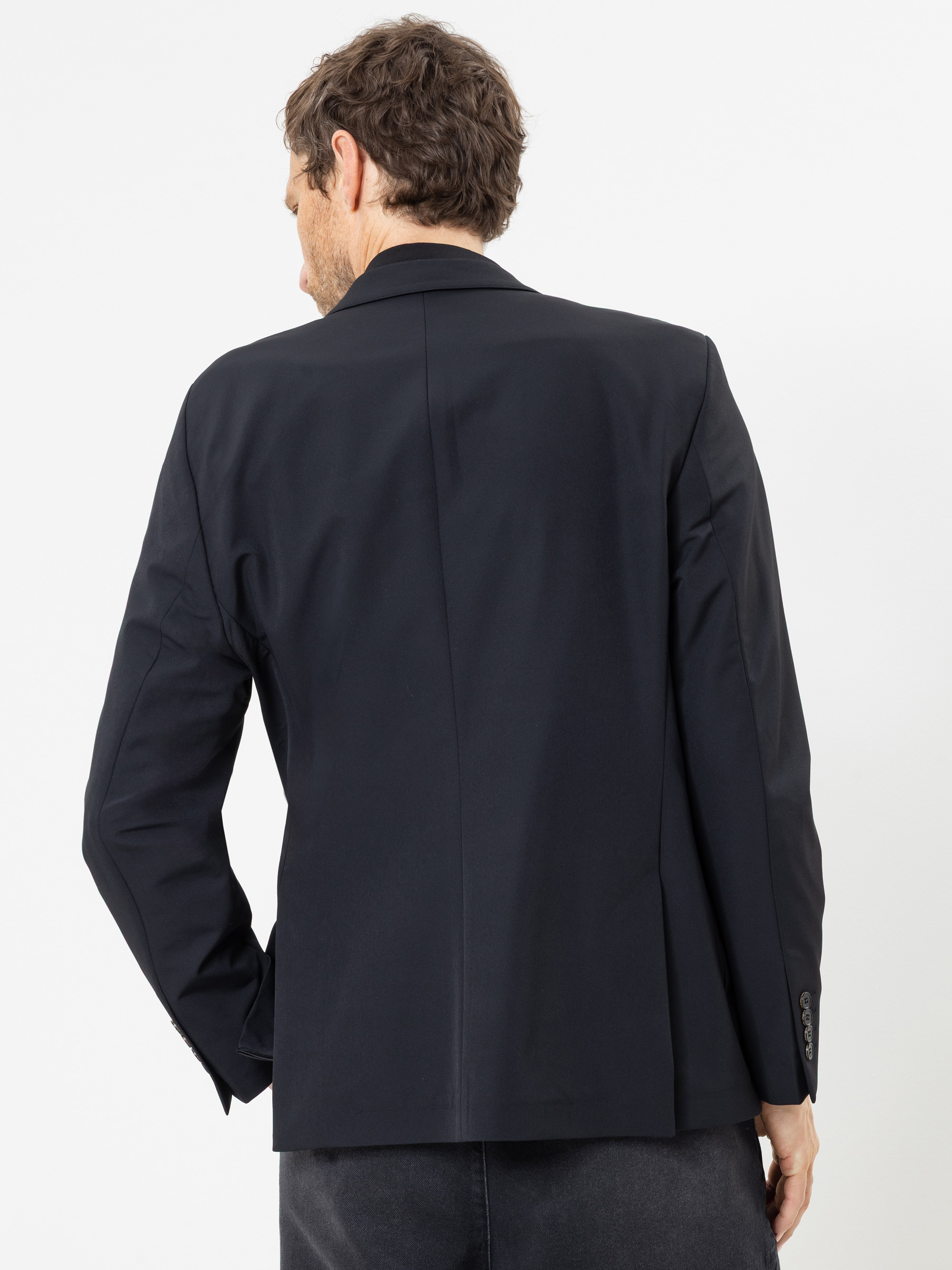 Ein Mann, der einen schwarzen Blazer und eine dunkle Hose trägt, steht mit dem Rücken zur Kamera vor einem schlichten weißen Hintergrund.