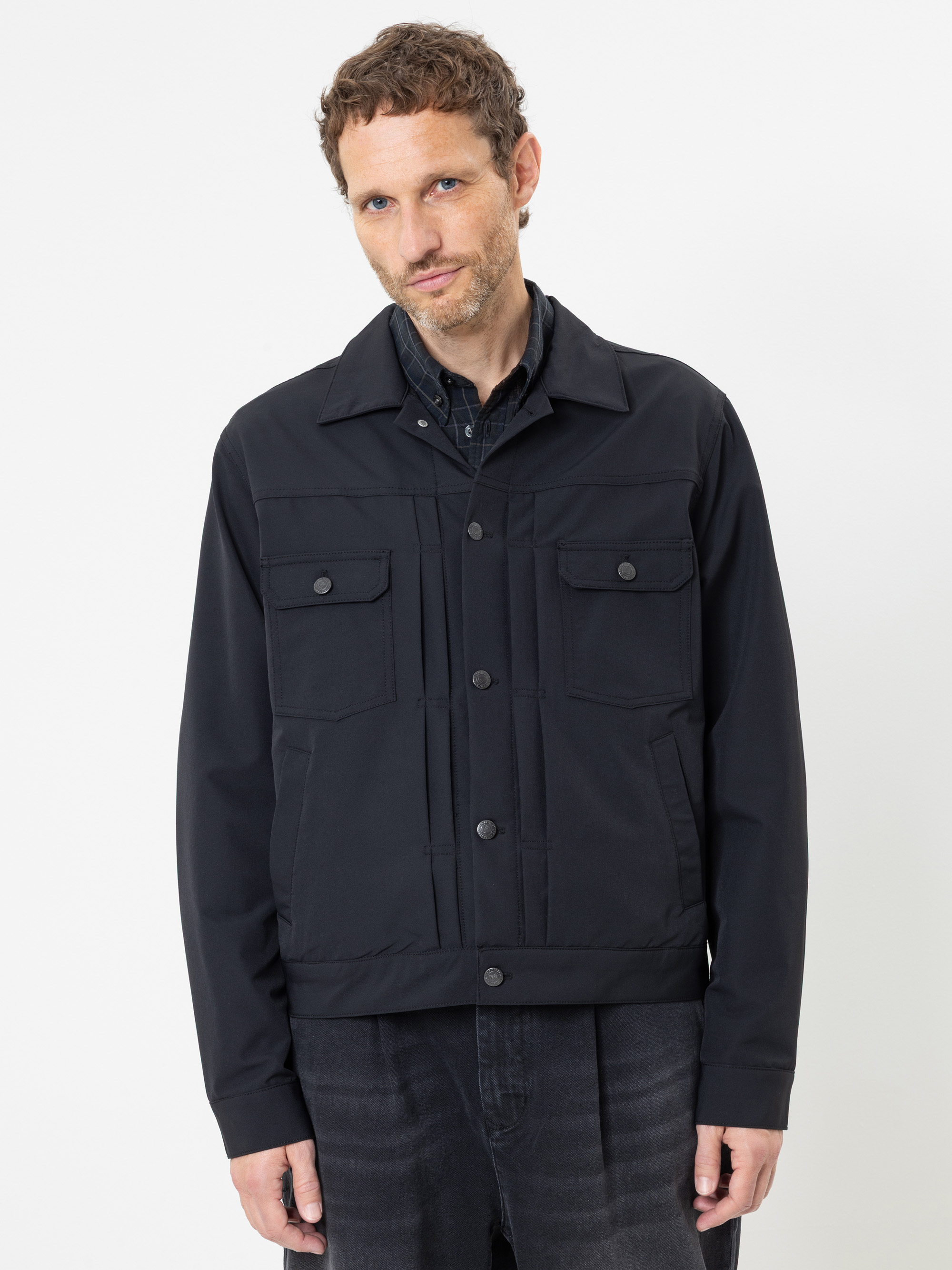Ein Mann mit kurzen braunen Haaren und Bart trägt eine schwarze Jacke mit Knöpfen und Pattentaschen über einem schwarzen Hemd und einer dunklen Hose und steht vor einem schlichten weißen Hintergrund.