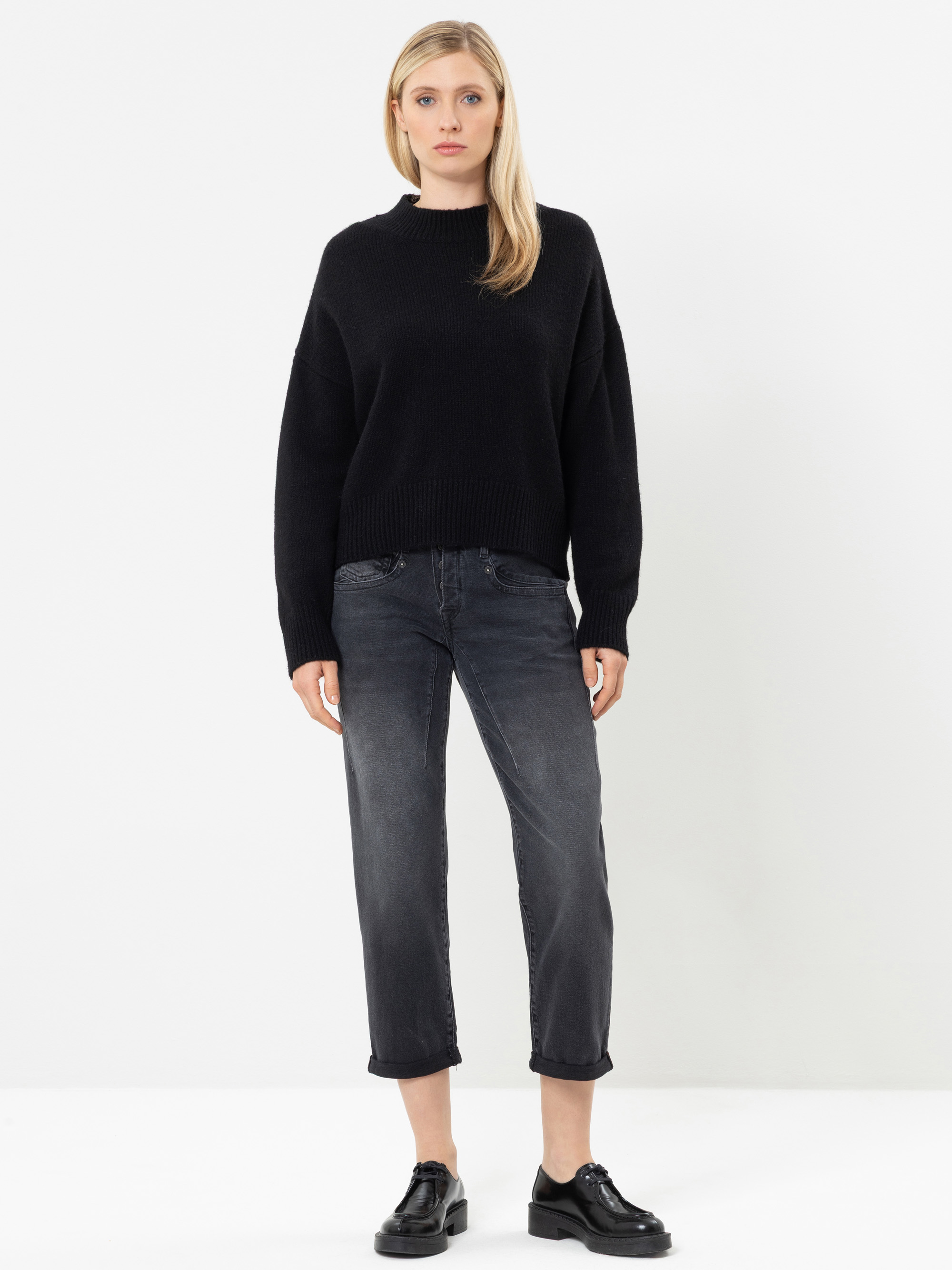 Vor einem schlichten weißen Hintergrund steht eine Frau, die einen schwarzen Pullover, ausgewaschene schwarze Jeans und schwarze Schuhe trägt. Sie hat glattes blondes Haar und einen ausdruckslosen Gesichtsausdruck; ihre Arme hängen entspannt an den Sei