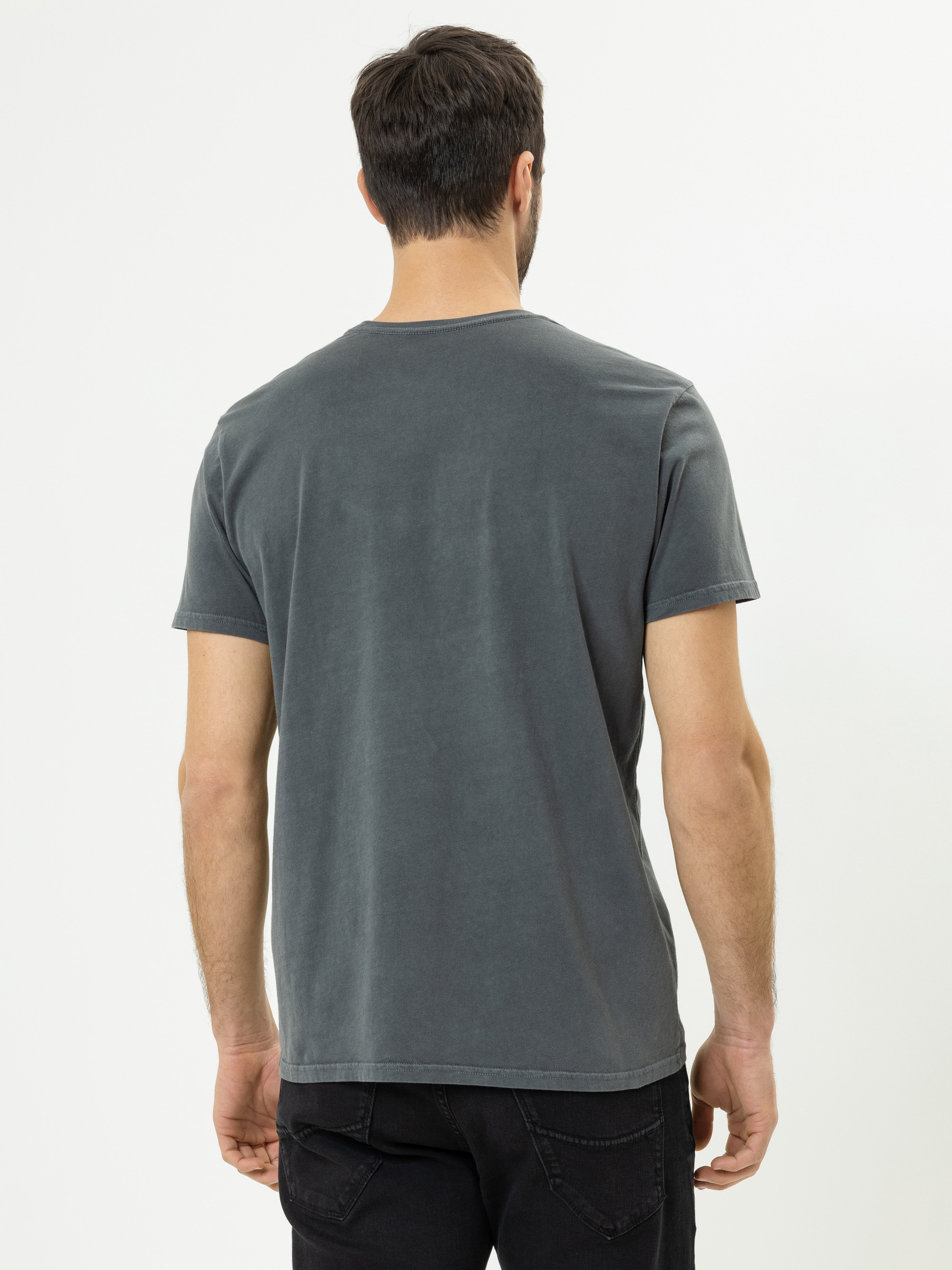 Ein Mann steht mit dem Rücken zur Kamera. Er trägt ein verblasstes, dunkelgraues, kurzärmeliges T-Shirt und schwarze Jeans vor einem schlichten weißen Hintergrund.