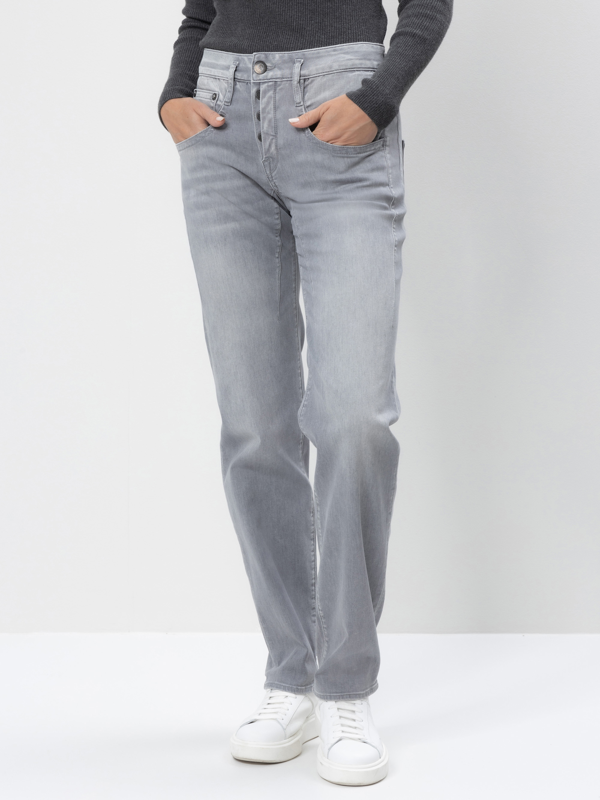 Eine Person, die ein dunkelgraues langärmeliges Oberteil, hellgraue Jeans mit geradem Bein und weiße Turnschuhe trägt, steht mit den Händen in den Taschen vor einem schlichten hellen Hintergrund.