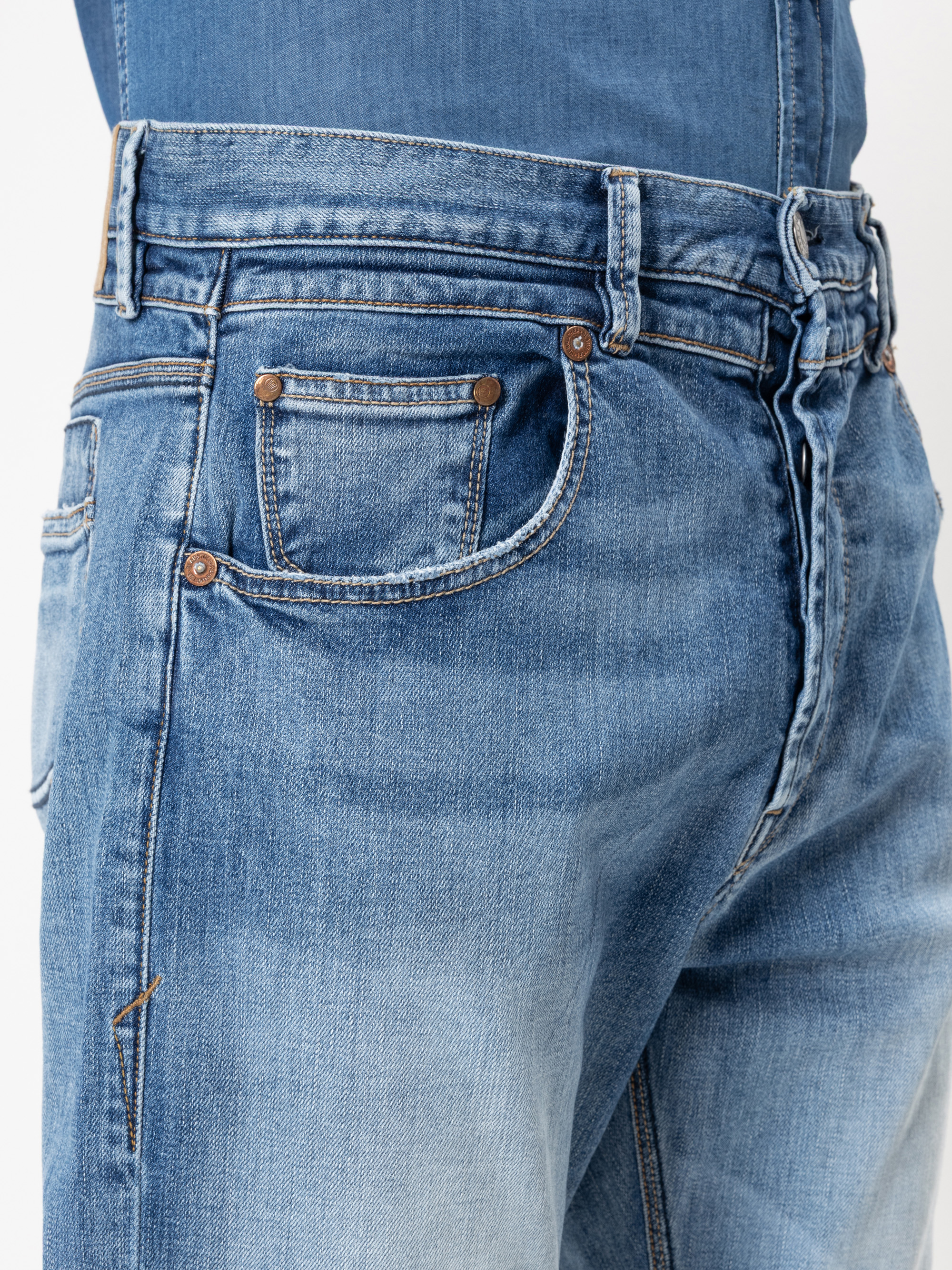 Nahaufnahme einer Person, die eine hellblaue Jeans und ein Jeanshemd trägt, wobei der Bund, die Gürtelschlaufen, die Tasche und die Metallnieten zu sehen sind. Die Jeans ist leicht verblasst und sieht abgenutzt aus.