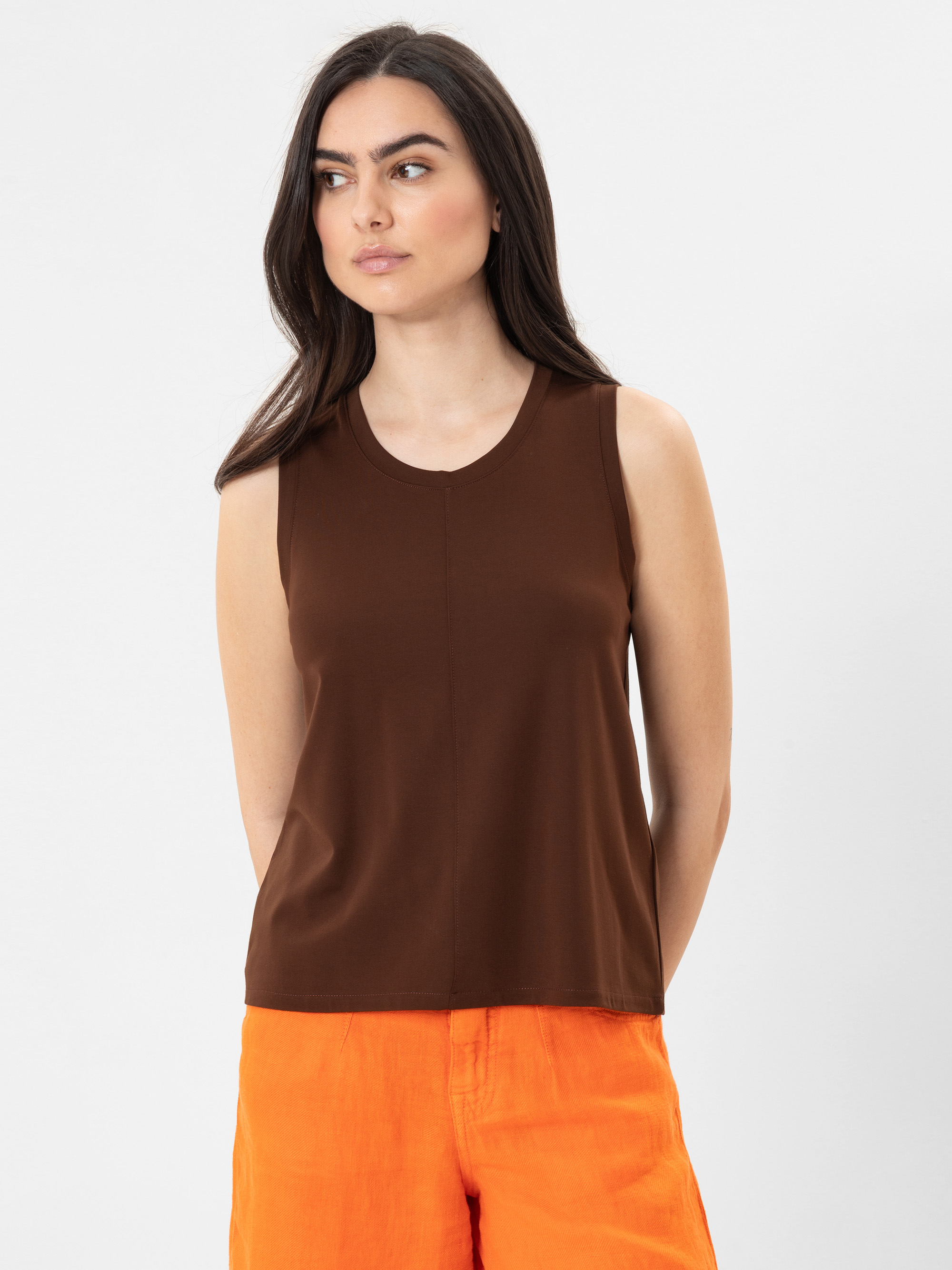 Eine Frau mit langen dunklen Haaren trägt ein ärmelloses braunes Oberteil und leuchtend orangefarbene Shorts. Sie steht vor einem schlichten weißen Hintergrund und schaut leicht nach links.