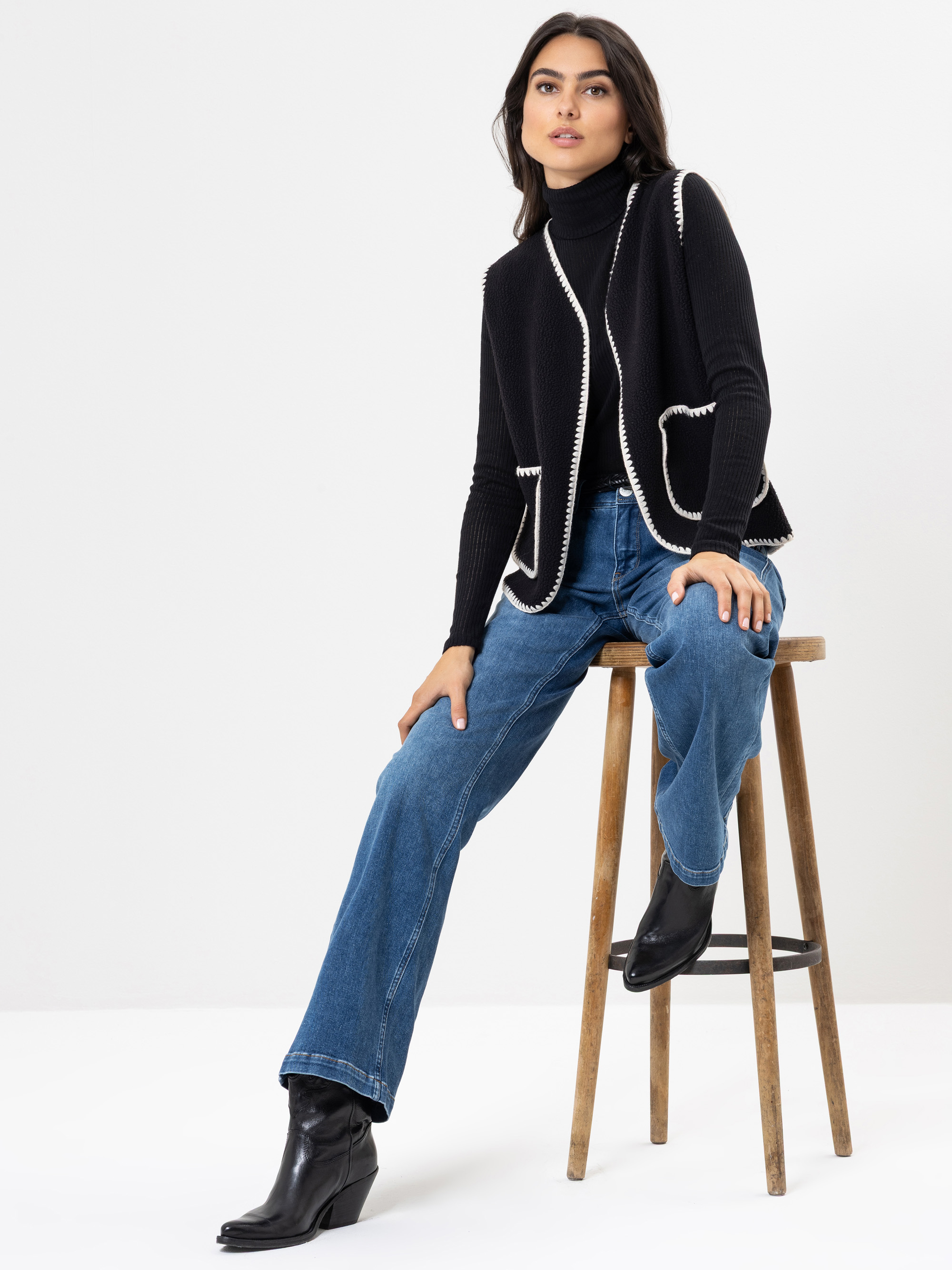 Eine Frau mit langen dunklen Haaren sitzt auf einem Holzschemel und trägt einen schwarzen Rollkragenpullover, eine schwarze Weste mit weißem Saum, blaue Jeans und schwarze Stiefeletten vor einem weißen Hintergrund.