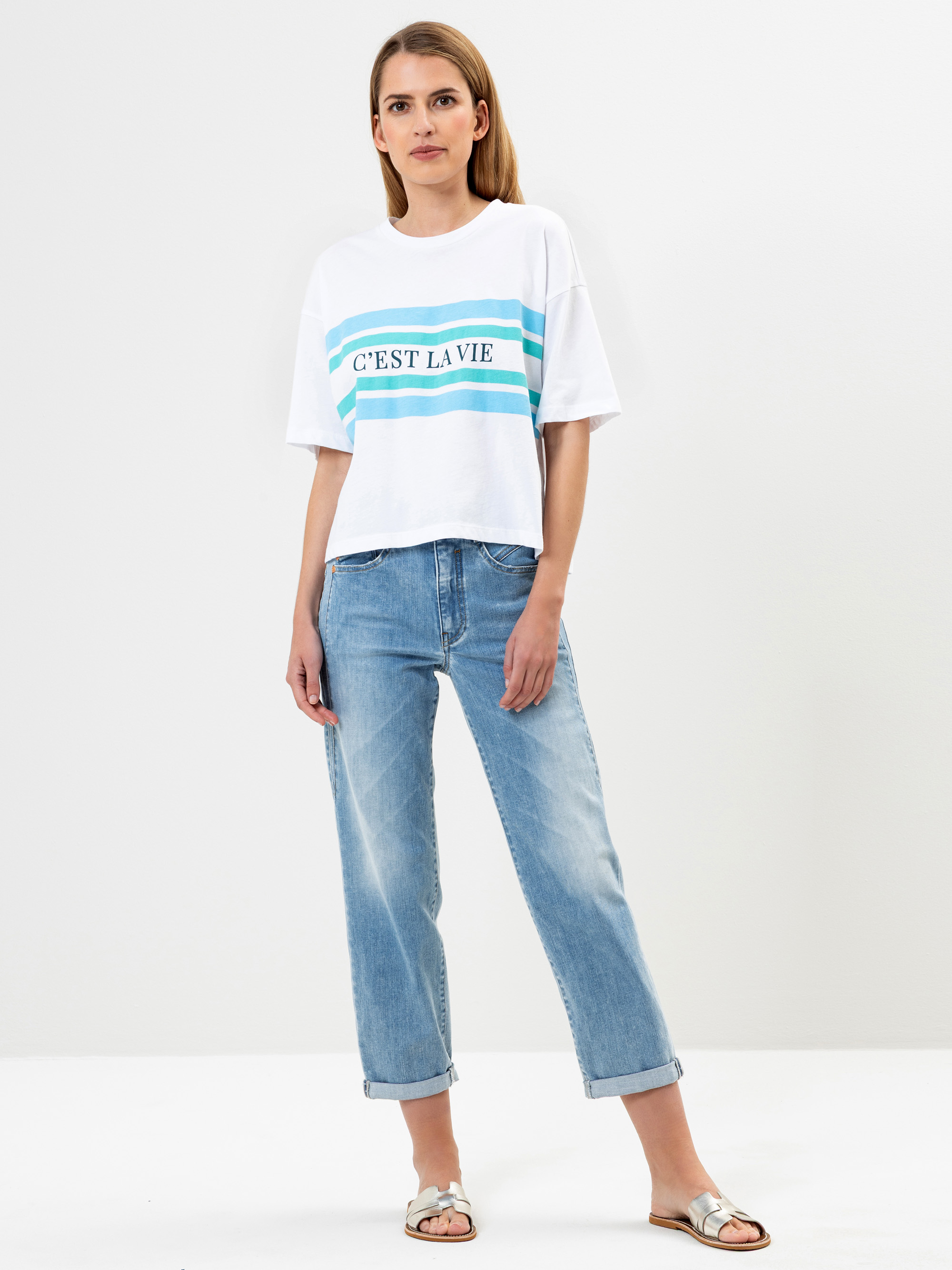 Eine Frau steht vor einem schlichten weißen Hintergrund und trägt ein weißes T-Shirt mit blauen Streifen und der Aufschrift C'EST LA VIE, blaue Jeans mit Bündchen und weiße Sandalen. Sie hat glattes, hellbraunes Haar und einen entspannten Gesichtsaus