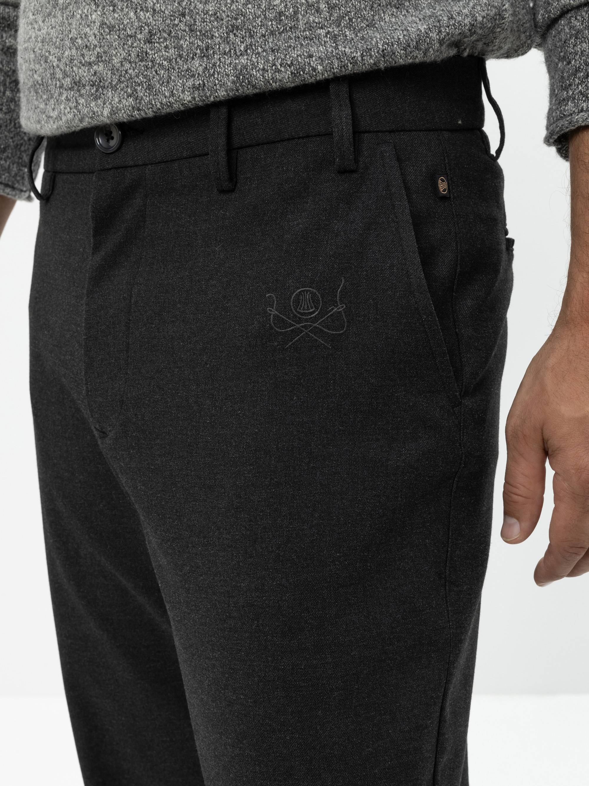 Eine Person trägt eine dunkelgraue maßgeschneiderte Hose mit einem kleinen gestickten Muster in der Nähe der Tasche und einen grauen Strickpullover, der von der Taille abwärts mit einer sichtbaren Hand gezeigt wird.