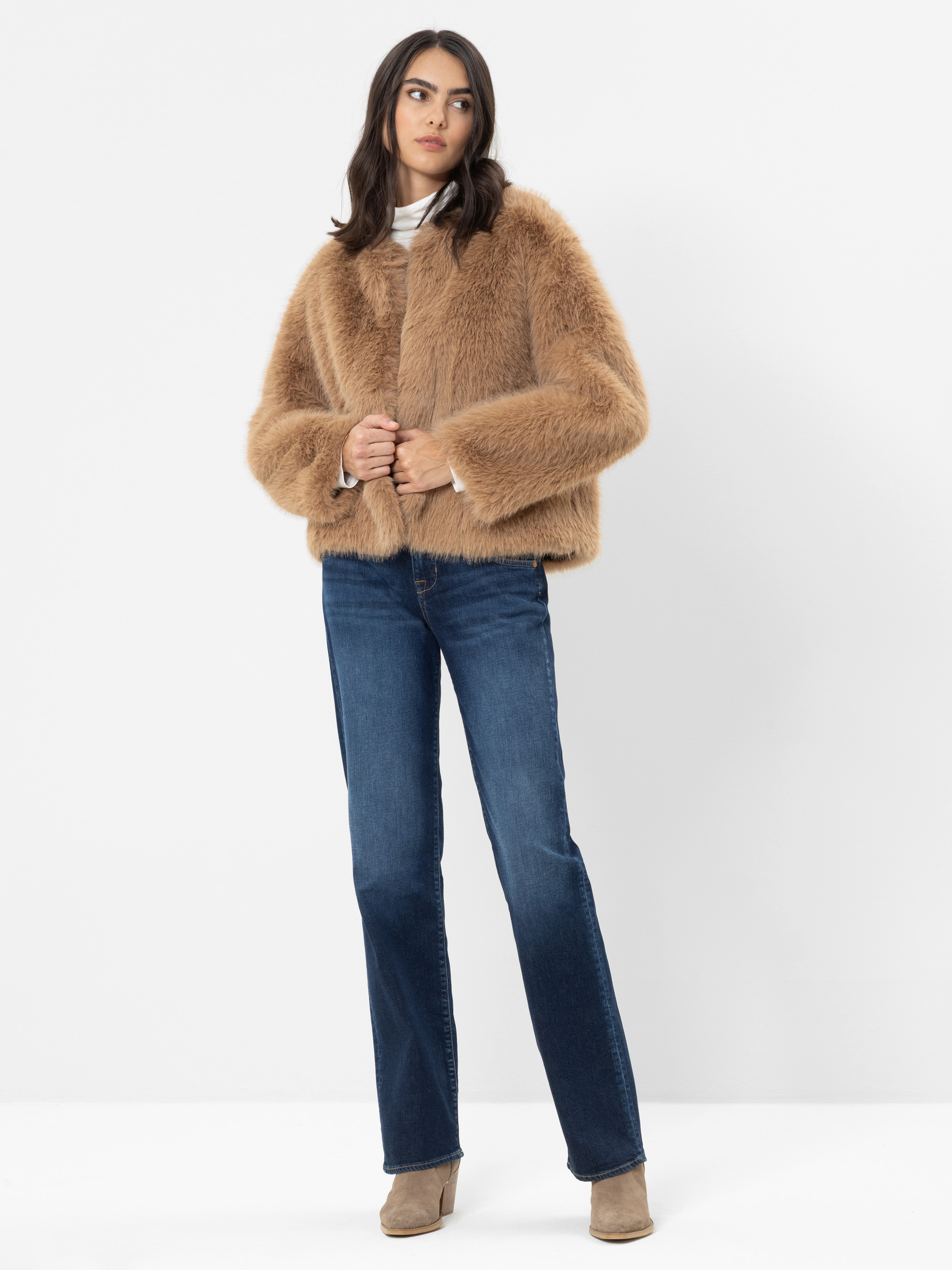 Eine Frau steht vor einem weißen Hintergrund, trägt eine hellbraune Kunstfelljacke, blaue Jeans, taupefarbene Stiefeletten und einen weißen Rollkragenpullover; sie blickt leicht zur Seite, während ihre Hände auf ihrer Jacke ruhen.