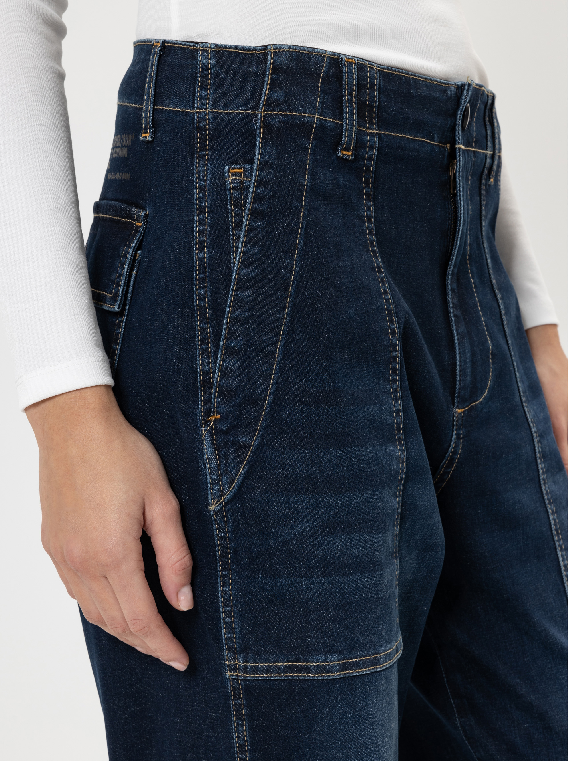 Eine Person trägt eine dunkelblaue Jeans mit hoher Taille und Kontrastnähten und ein weißes langärmeliges Hemd. Der Schwerpunkt liegt auf den Taschen und Nähten der Jeans, während die Hand der Person an ihrer Seite ruht.