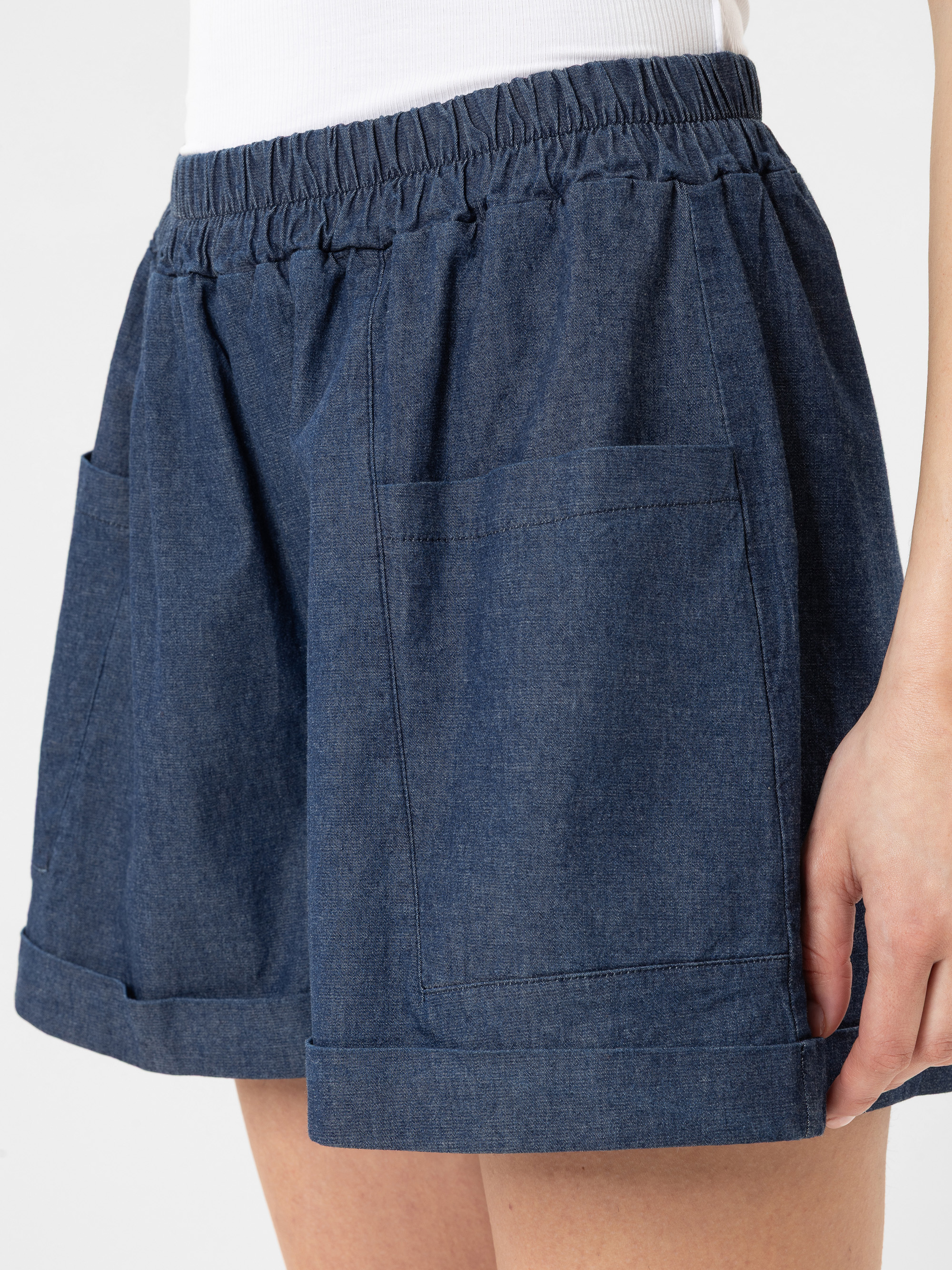 Eine Person trägt dunkelblaue Jeansshorts mit elastischem Bund und großen Vordertaschen, kombiniert mit einem weißen Oberteil. Die Shorts haben eine lockere Passform und gerollte Bündchen.