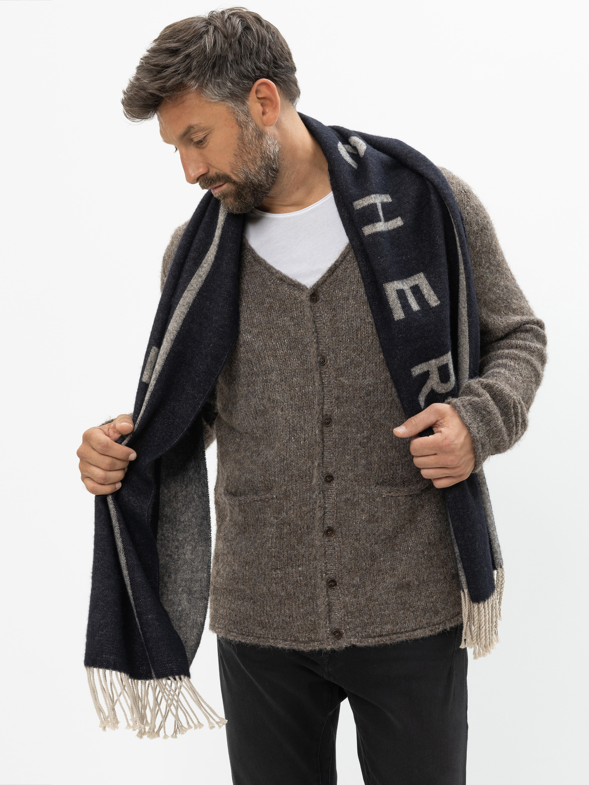 Ein Mann mit kurzen braunen Haaren und einem Bart trägt eine braune Strickjacke, ein weißes T-Shirt und eine schwarze Hose. Er blickt nach unten, während er einen langen, dunklen Schal mit hellen Fransen und großen Buchstaben darauf zurechtrückt. Der