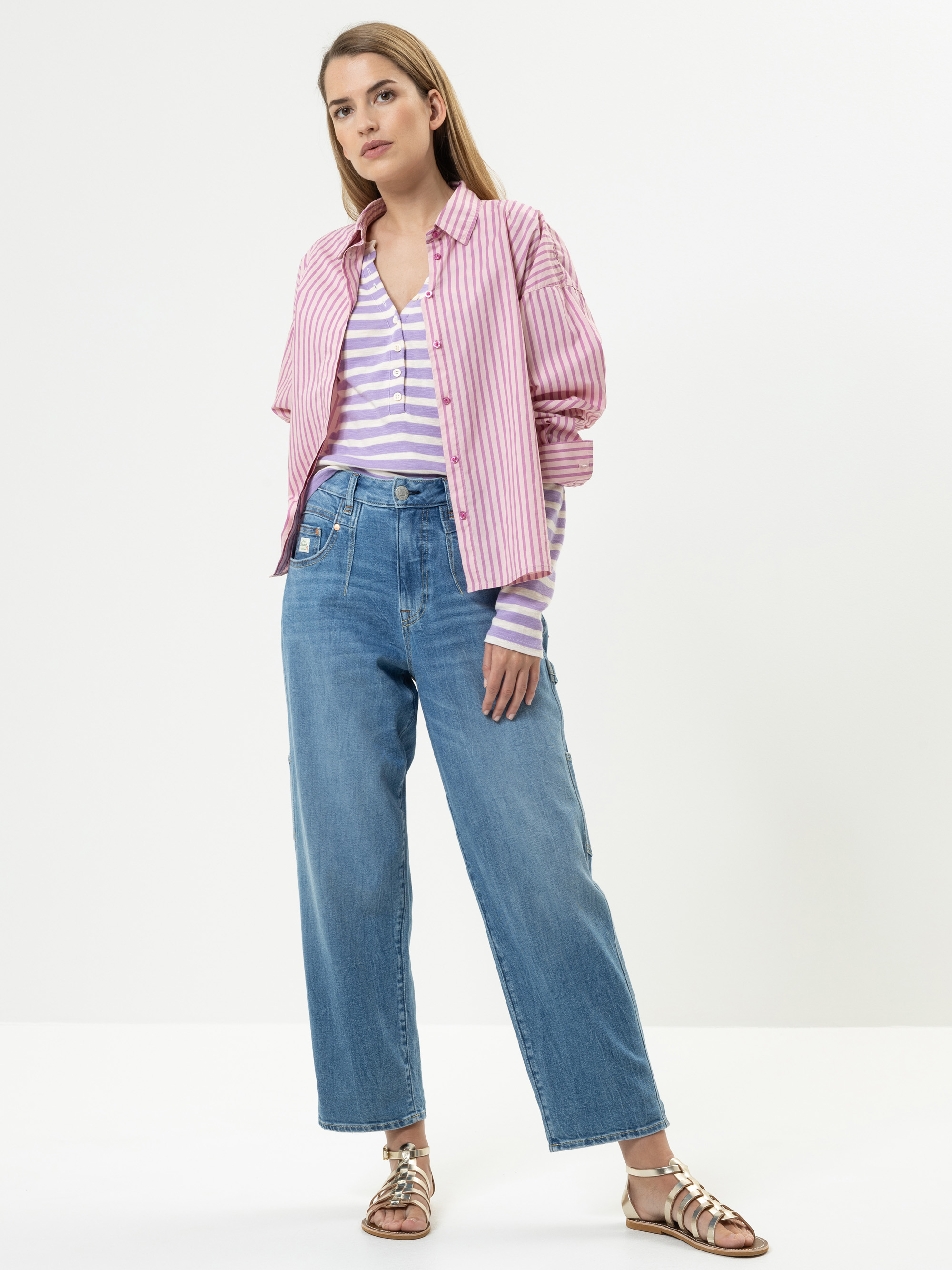 Eine Frau steht vor einem weißen Hintergrund und trägt ein rosa gestreiftes Hemd über einem lila-weiß gestreiften Top, blaue High-Waisted-Jeans und Metallic-Sandalen. Sie hat langes, glattes Haar und eine entspannte Pose.