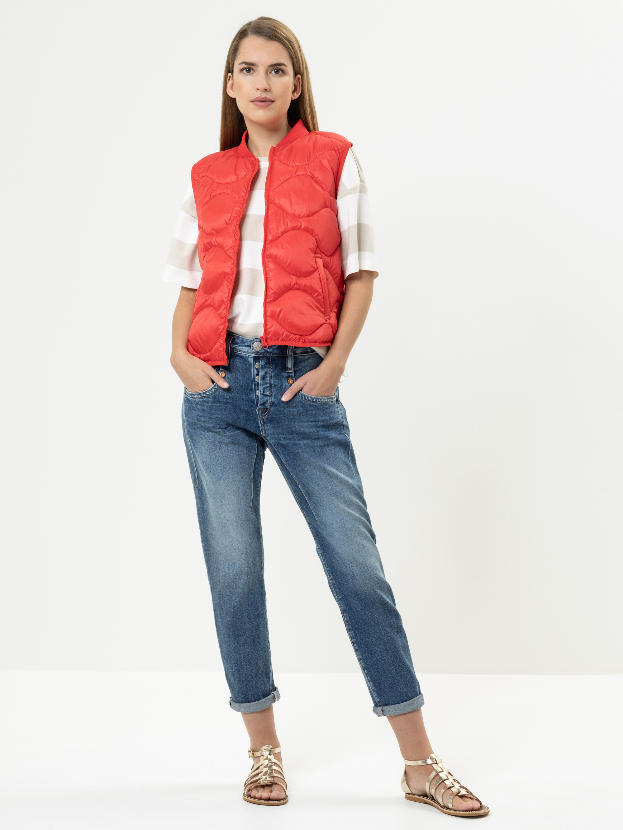 Eine Frau steht selbstbewusst in einer roten Pufferweste über einem weiß-beige gestreiften Hemd, blauen Jeans mit Bündchen und goldenen Sandalen vor einem schlichten weißen Hintergrund.