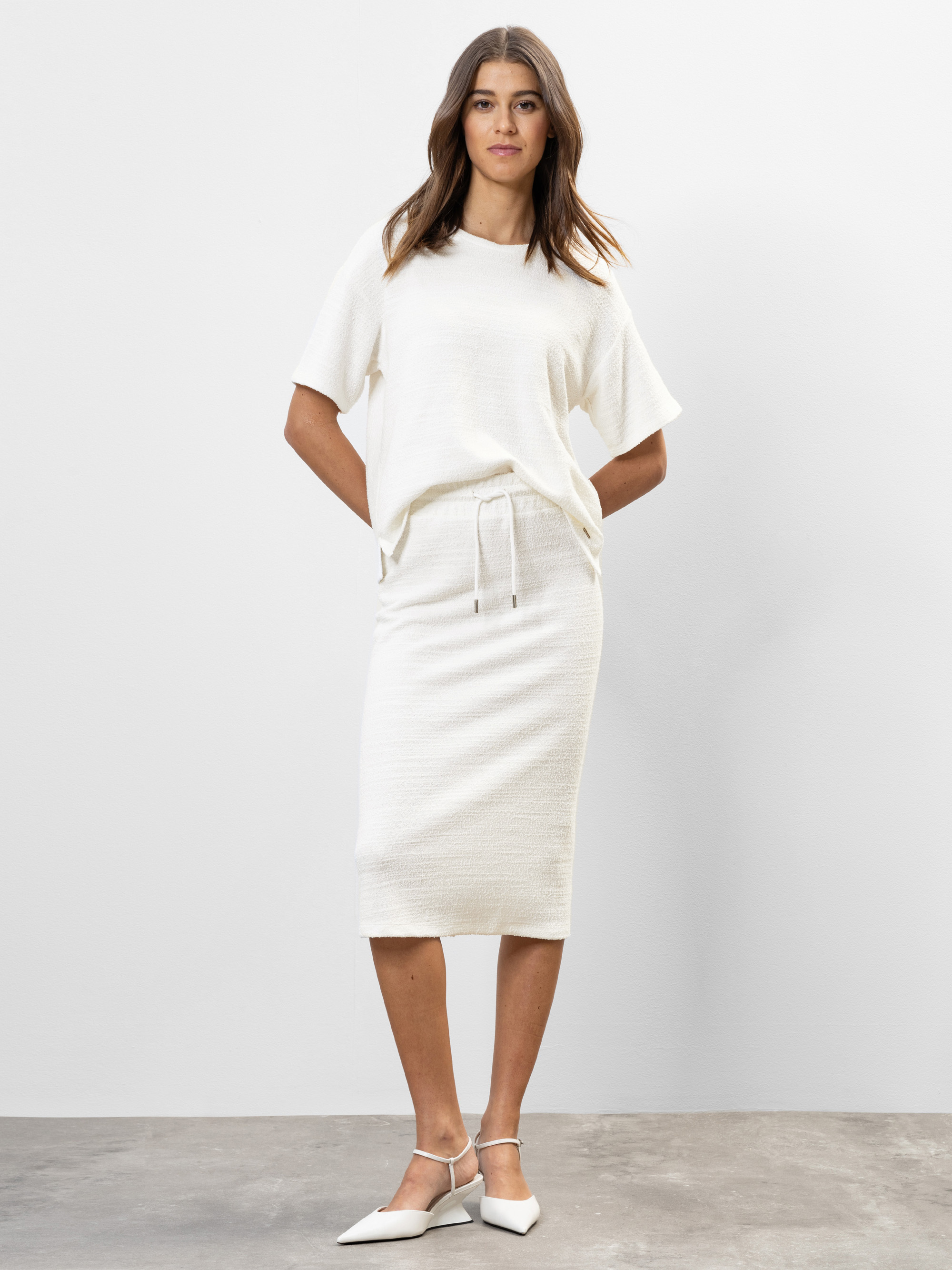 Eine Frau steht vor einem schlichten weißen Hintergrund und trägt ein kurzärmeliges, knielanges weißes Strickkleid mit Tunnelzug und weißen spitzen Schuhen. Ihre Hände sind hinter dem Rücken verschränkt, und sie hat lockeres, gewelltes Haar.