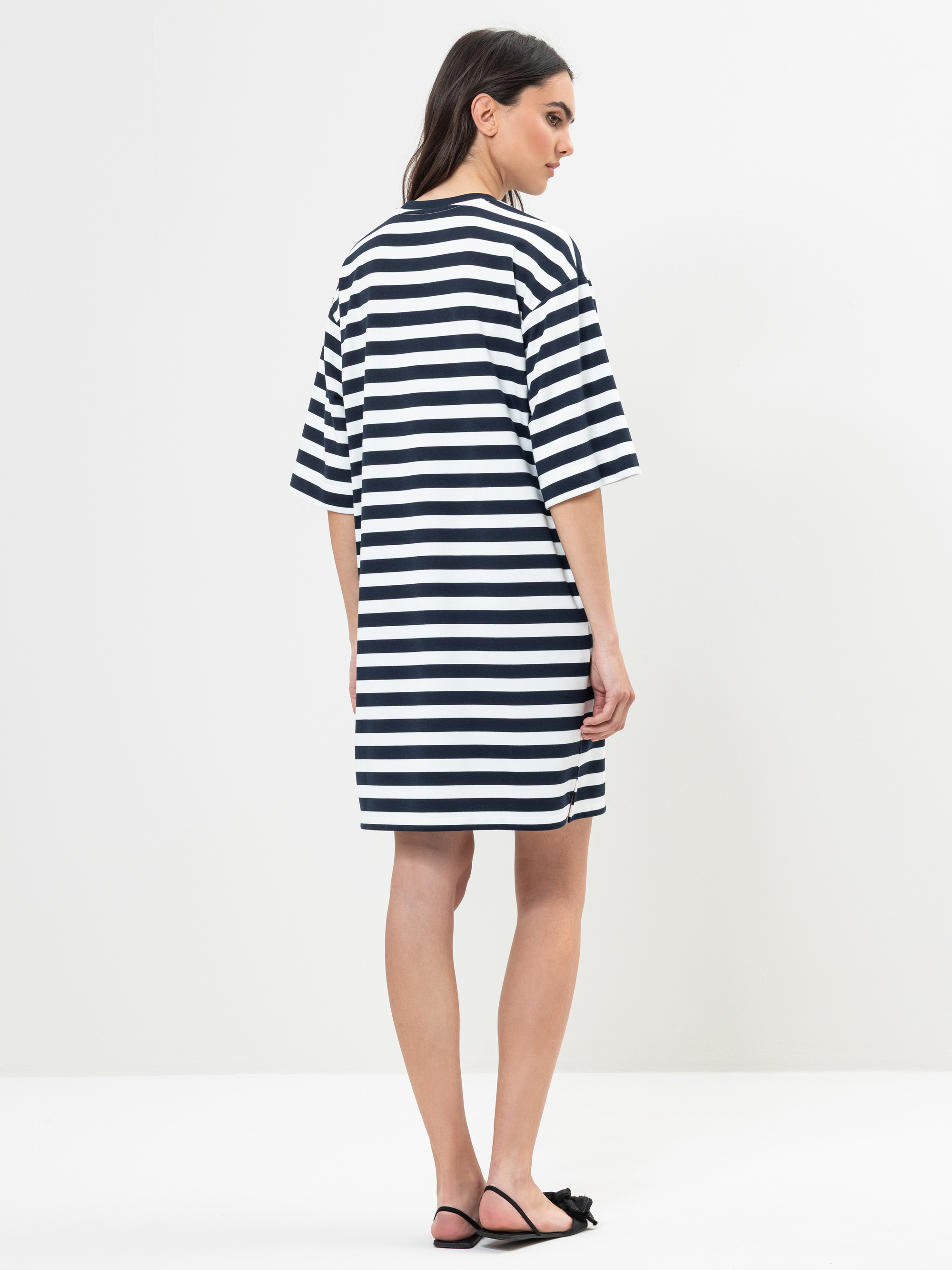 Eine Frau steht mit dem Gesicht zur Wand und trägt ein lockeres, schwarz-weißes, horizontal gestreiftes Kleid mit kurzen Ärmeln. Sie hat langes dunkles Haar und trägt schwarze Sandalen. Der Hintergrund ist schlicht und weiß.