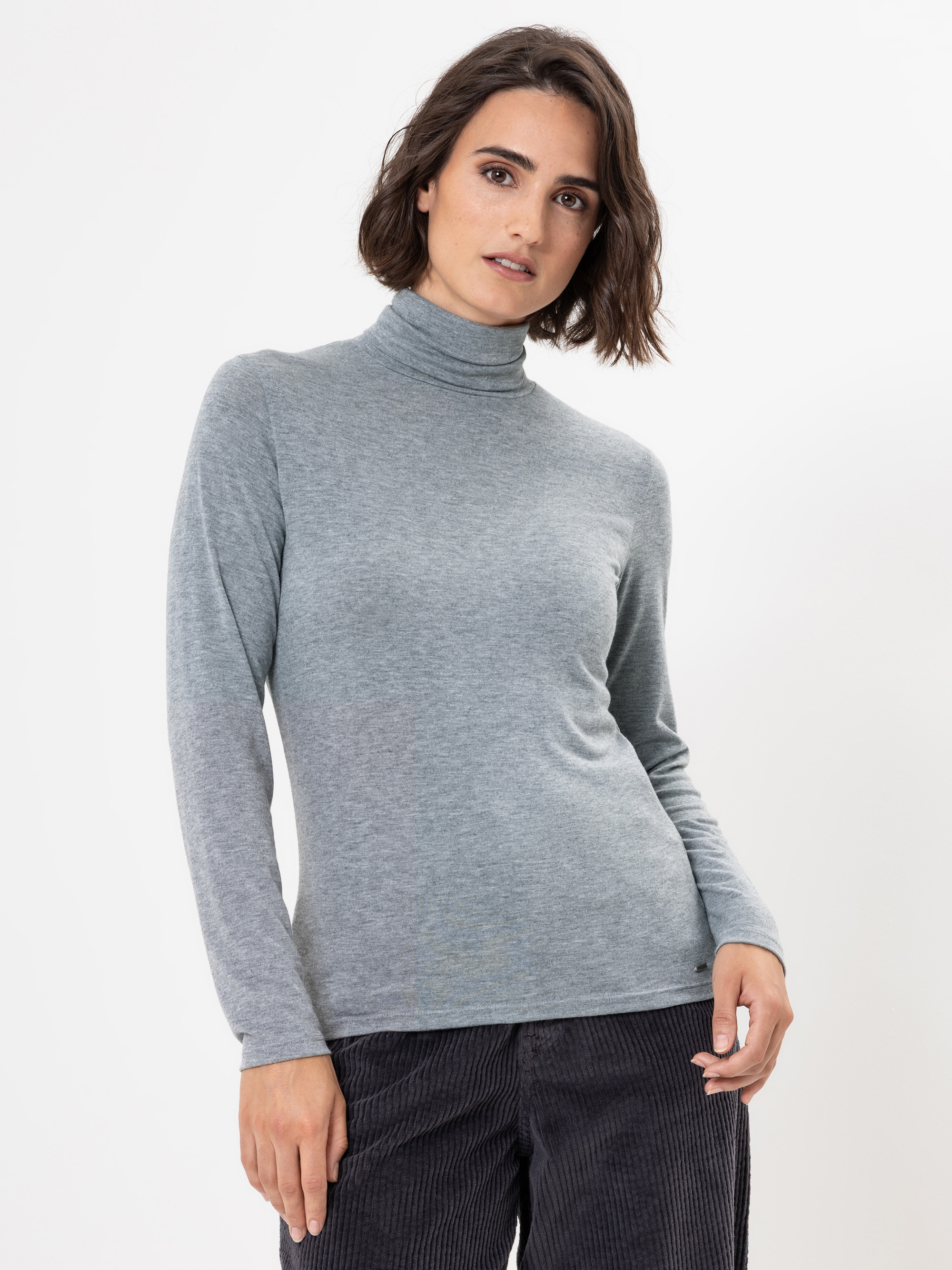 Eine Frau mit kurzen braunen Haaren, die einen hellgrauen Rollkragenpullover und eine dunkle Hose trägt, steht vor einem schlichten weißen Hintergrund und schaut selbstbewusst in die Kamera.