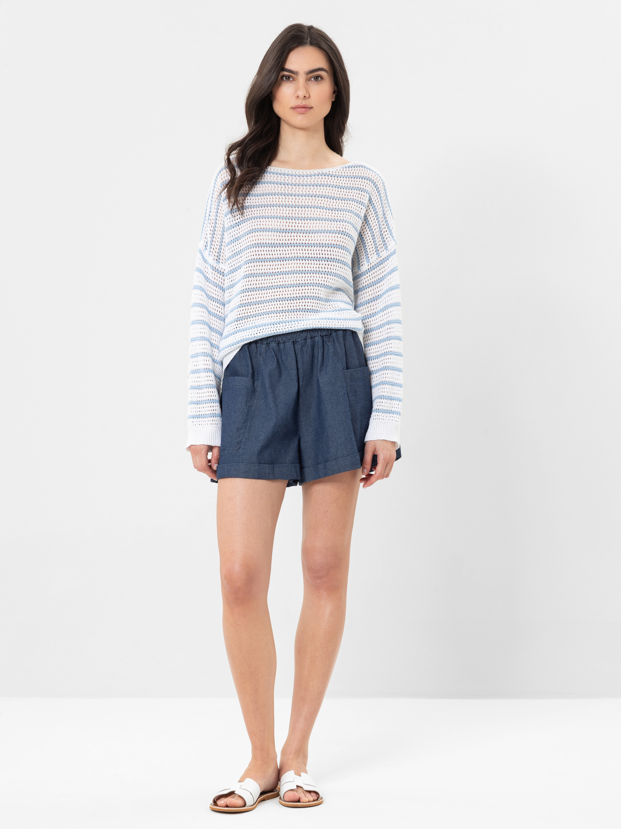 Eine Frau steht vor einem schlichten Hintergrund und trägt einen weiß-hellblau gestreiften, langärmeligen Pullover, locker sitzende Jeans-Shorts und weiße Slide-Sandalen. Sie hat langes braunes Haar und einen neutralen Gesichtsausdruck.