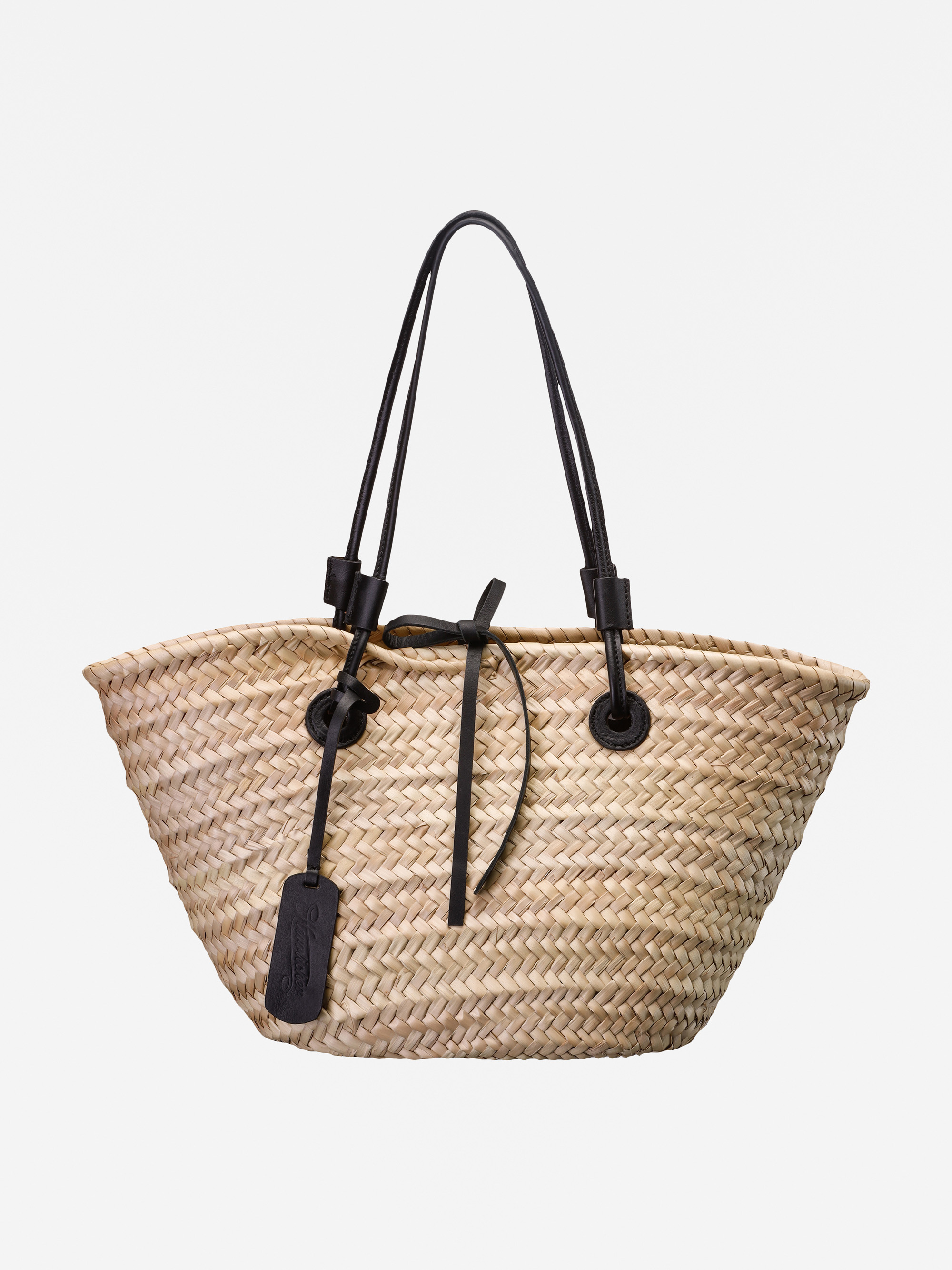 Eine Tragetasche aus gewebtem Stroh mit zwei langen schwarzen Ledergriffen und einem schwarzen Lederanhänger, fotografiert vor einem schlichten weißen Hintergrund.