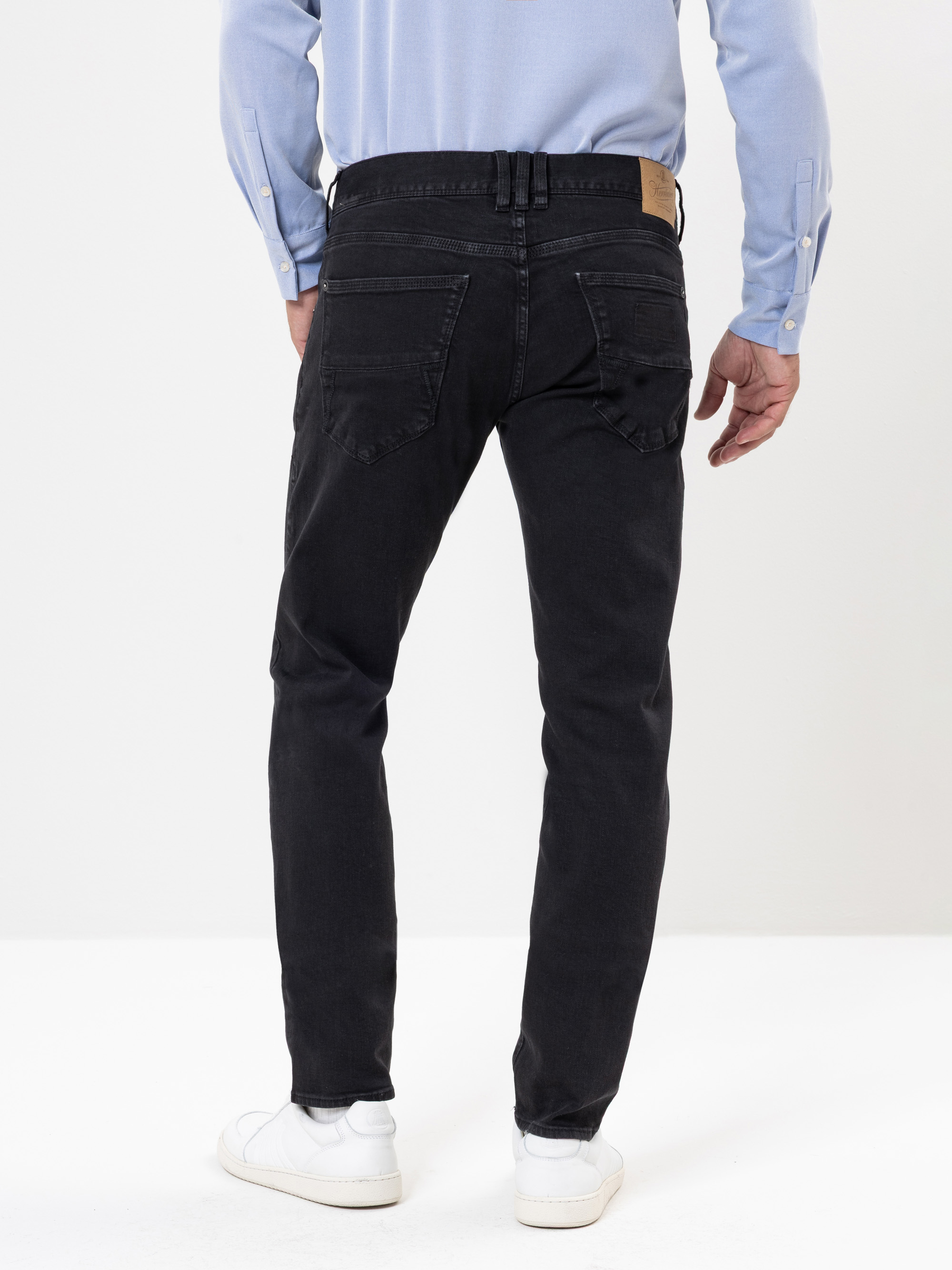 Eine Person, die ein hellblaues langärmeliges Hemd, schwarze Jeans und weiße Turnschuhe trägt, steht mit dem Gesicht zur Kamera vor einem einfarbig weißen Hintergrund.