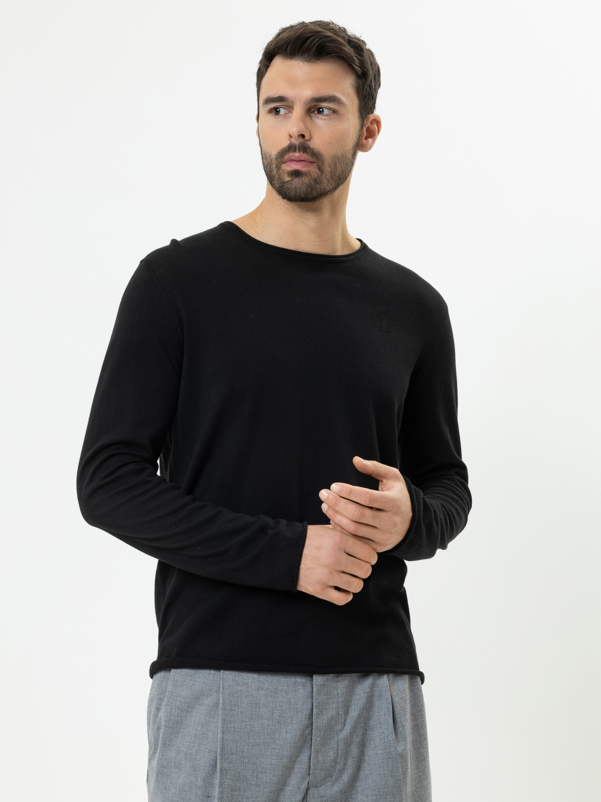 Ein Mann mit kurzen dunklen Haaren und einem Bart trägt ein schwarzes langärmeliges Hemd und eine hellgraue Hose und steht vor einem schlichten weißen Hintergrund. Er blickt leicht nach links und hat einen neutralen Blick.