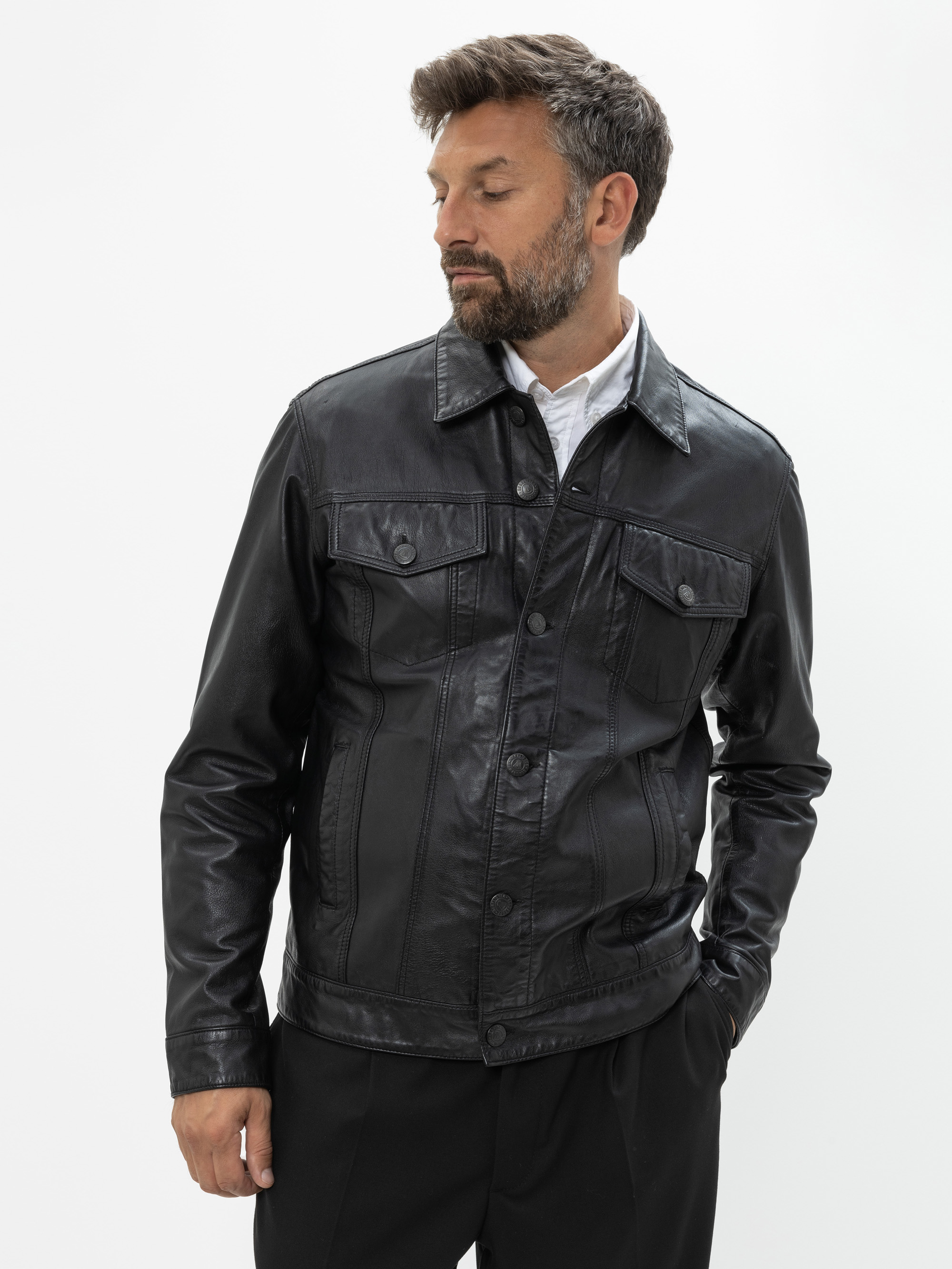 Ein Mann mit kurzen, dunklen Haaren und einem Bart trägt eine schwarze Lederjacke über einem weißen Hemd und einer schwarzen Hose. Er steht vor einem schlichten weißen Hintergrund und blickt leicht nach links, eine Hand in der Tasche.