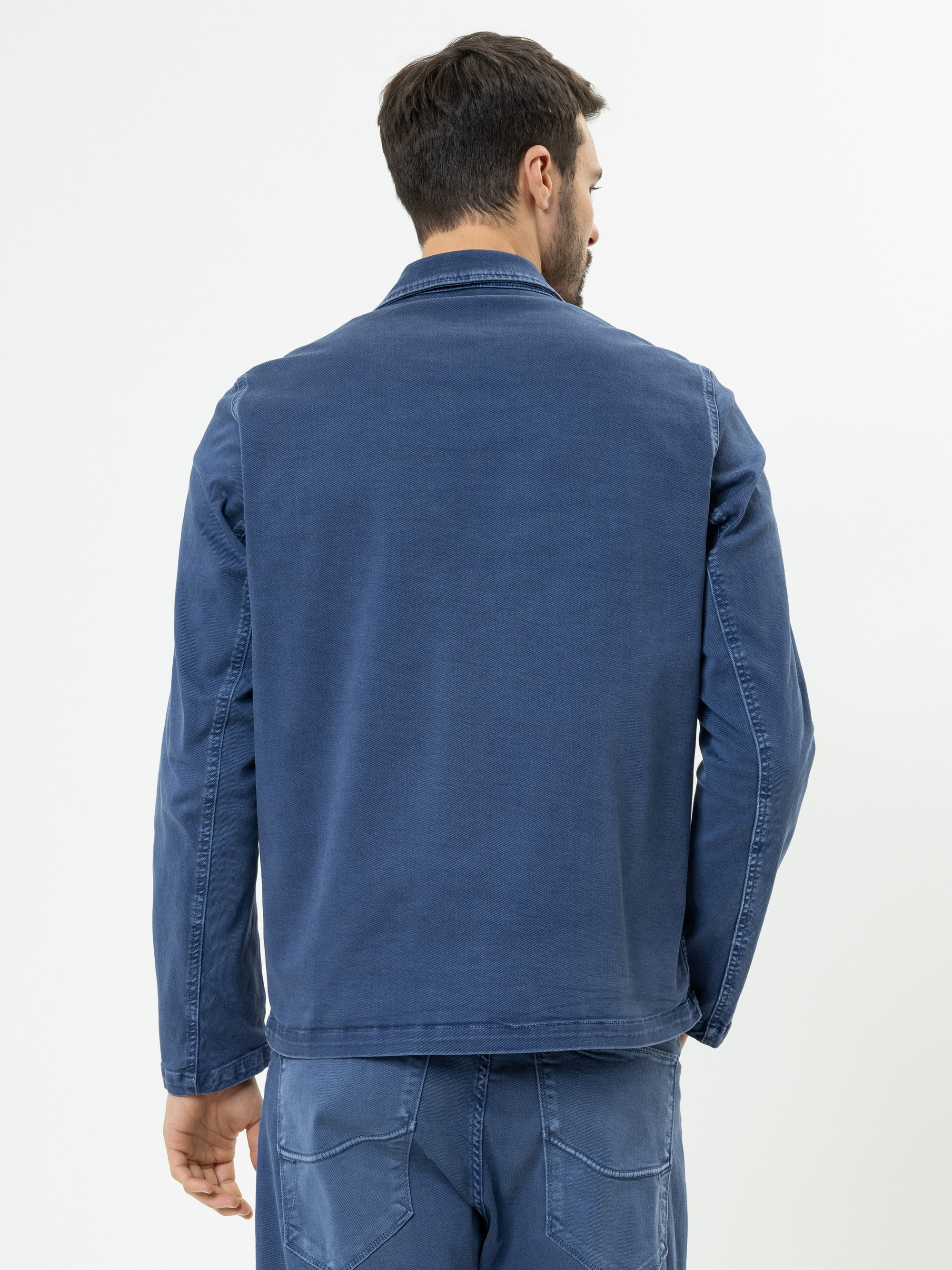 Ein Mann steht mit dem Rücken zur Kamera, trägt eine blaue Jeansjacke und eine dazu passende blaue Jeans vor einem schlichten weißen Hintergrund.