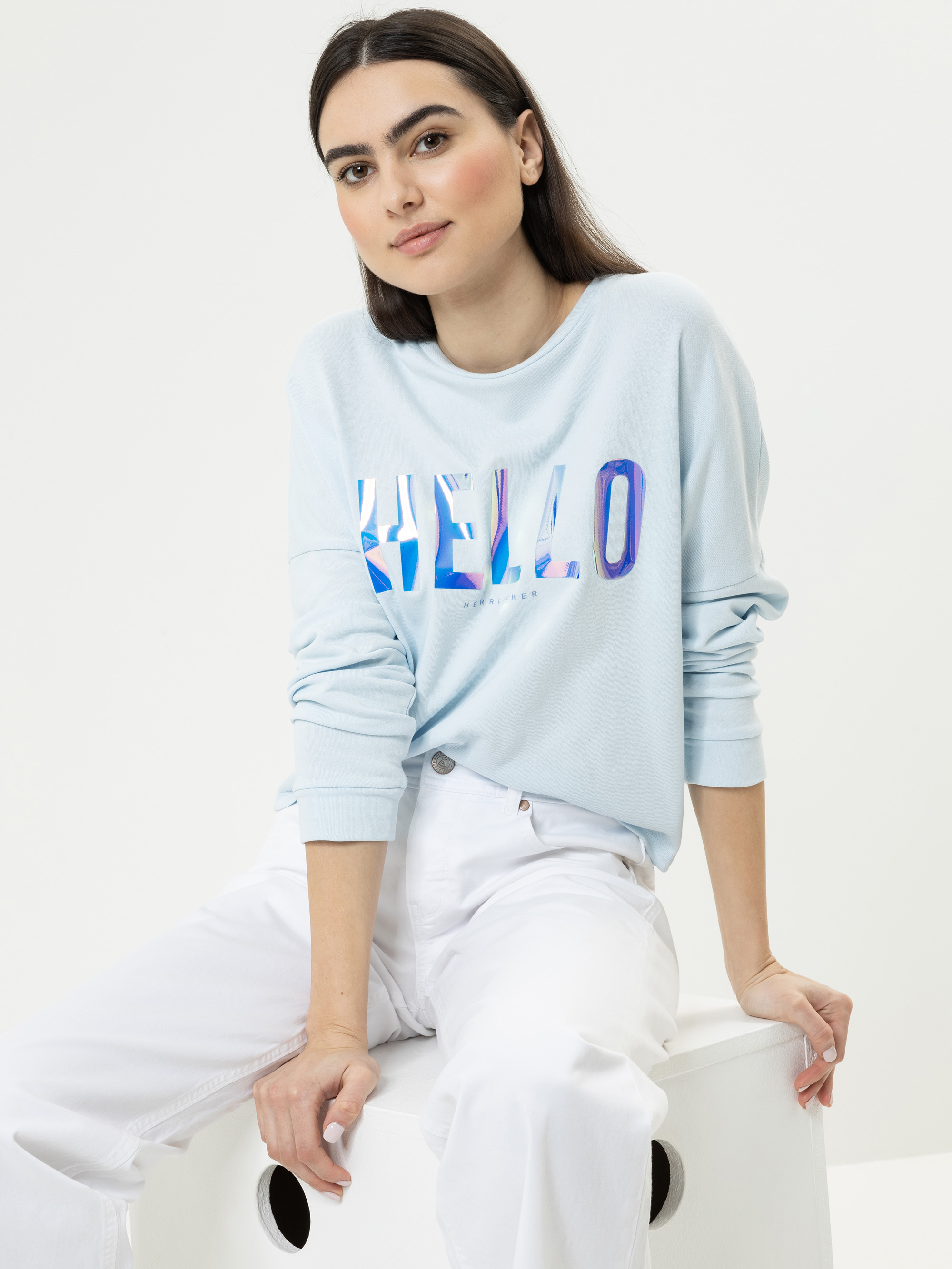 Eine Frau mit langen dunklen Haaren sitzt auf einem weißen Würfel, trägt ein hellblaues Sweatshirt mit dem Wort HELLO in bunten Buchstaben und eine weiße Hose vor einem schlichten weißen Hintergrund.