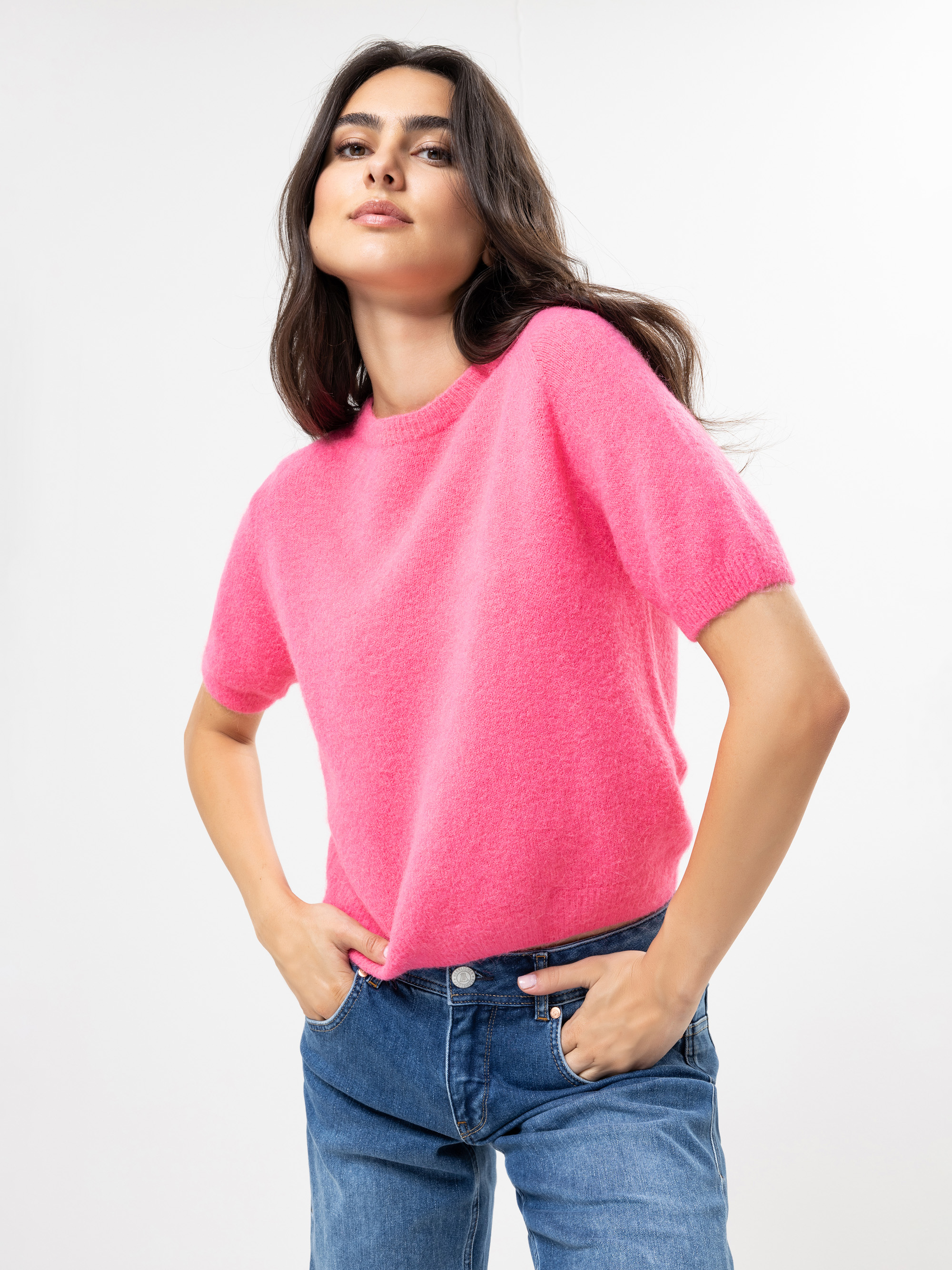 Eine Frau mit langen dunklen Haaren trägt einen hellrosa kurzärmeligen Pullover und blaue Jeans und steht mit den Händen in den Taschen vor einem schlichten weißen Hintergrund.
