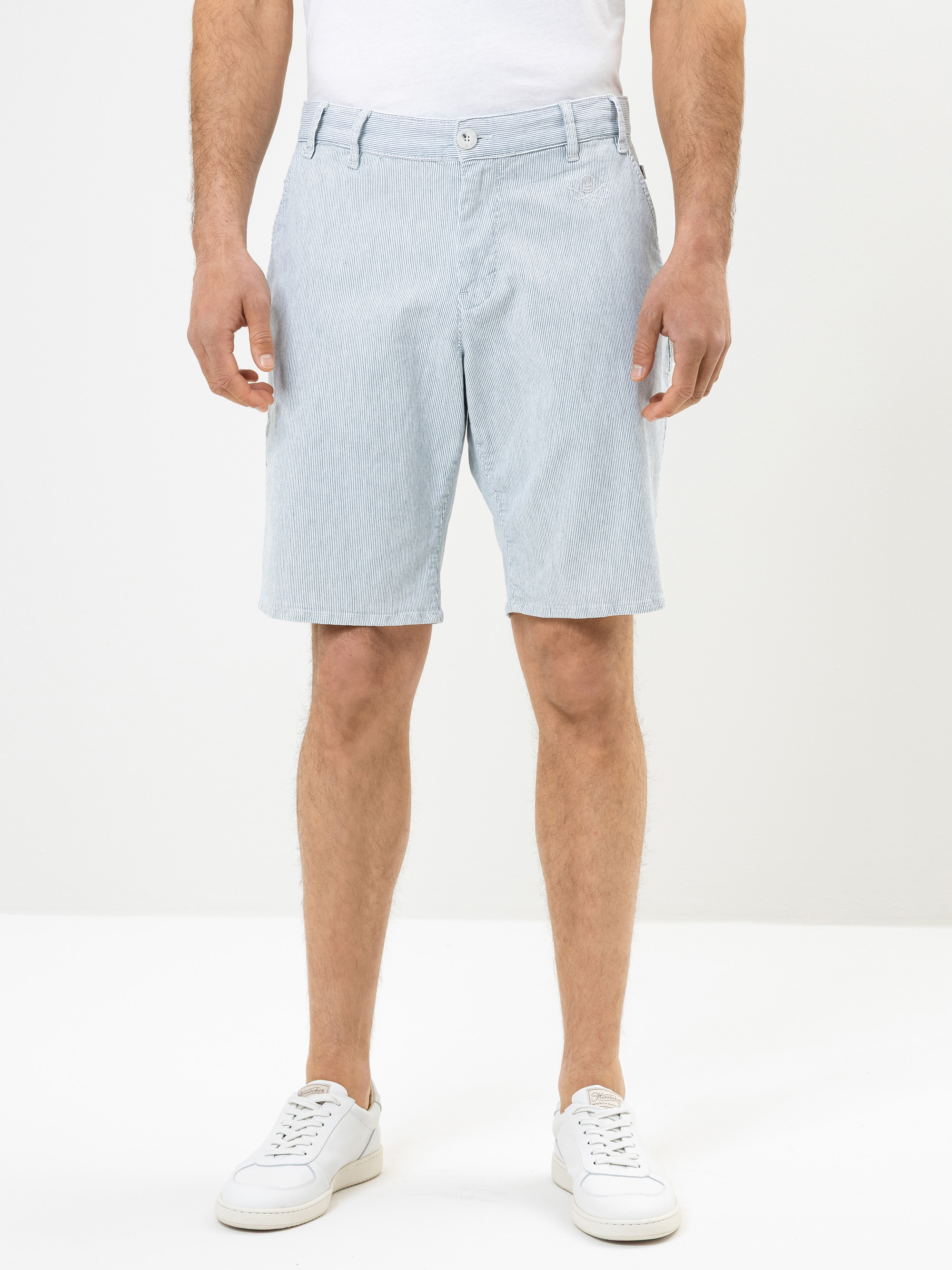 Ein Mann in hellblauen, knielangen Shorts, einem weißen T-Shirt und weißen Turnschuhen steht vor einem schlichten weißen Hintergrund. Seine Arme liegen entspannt an den Seiten, und nur sein Körper ist von den Schultern abwärts zu sehen.