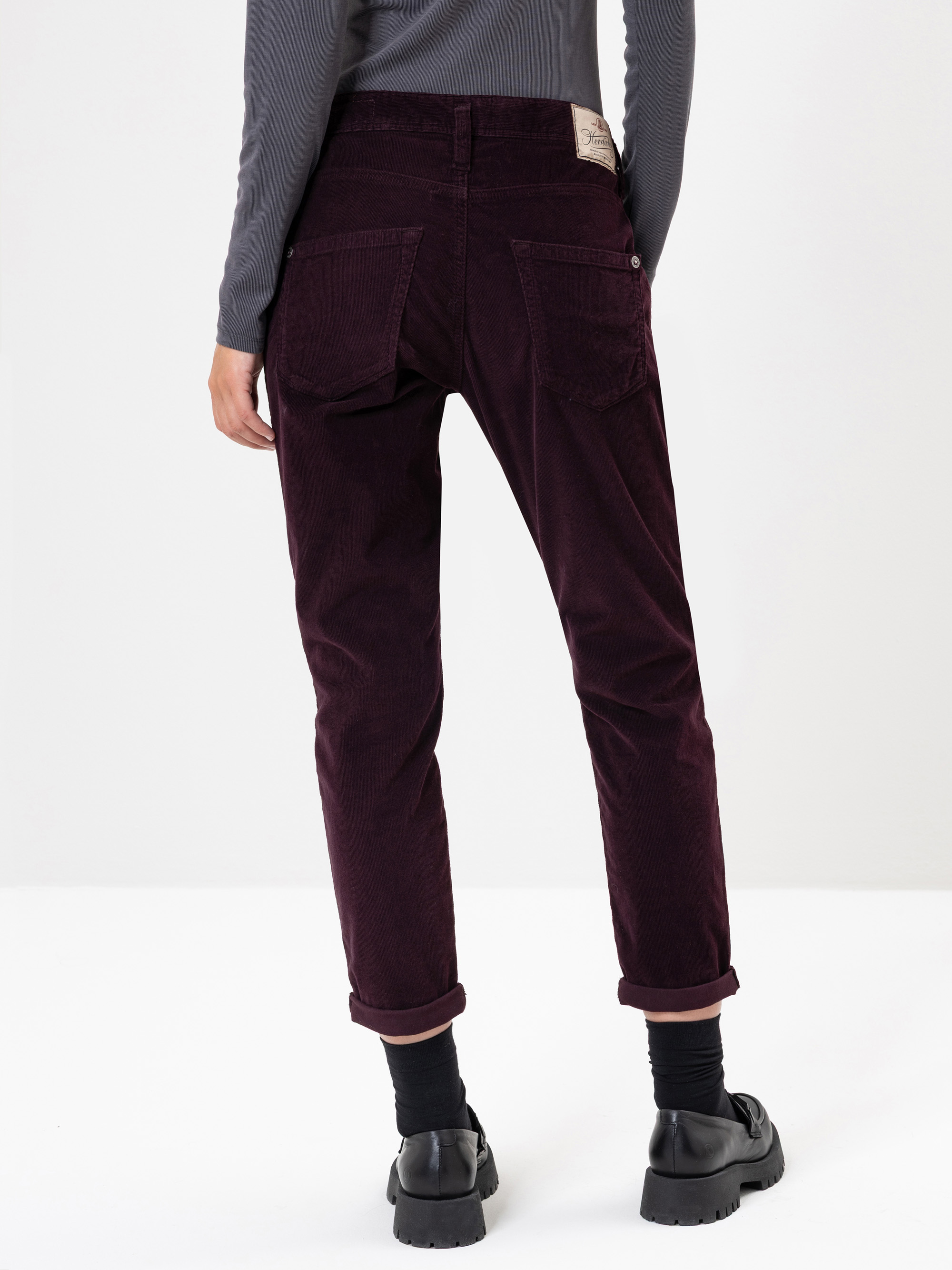 Eine Person steht mit dem Gesicht nach hinten und trägt eine dunkelbraune Cordhose, ein graues langärmeliges Oberteil, schwarze Socken und schwarze Schuhe. Die Hose hat aufgerollte Manschetten und sichtbare Gesäßtaschen. Der Hintergrund ist einfarbig