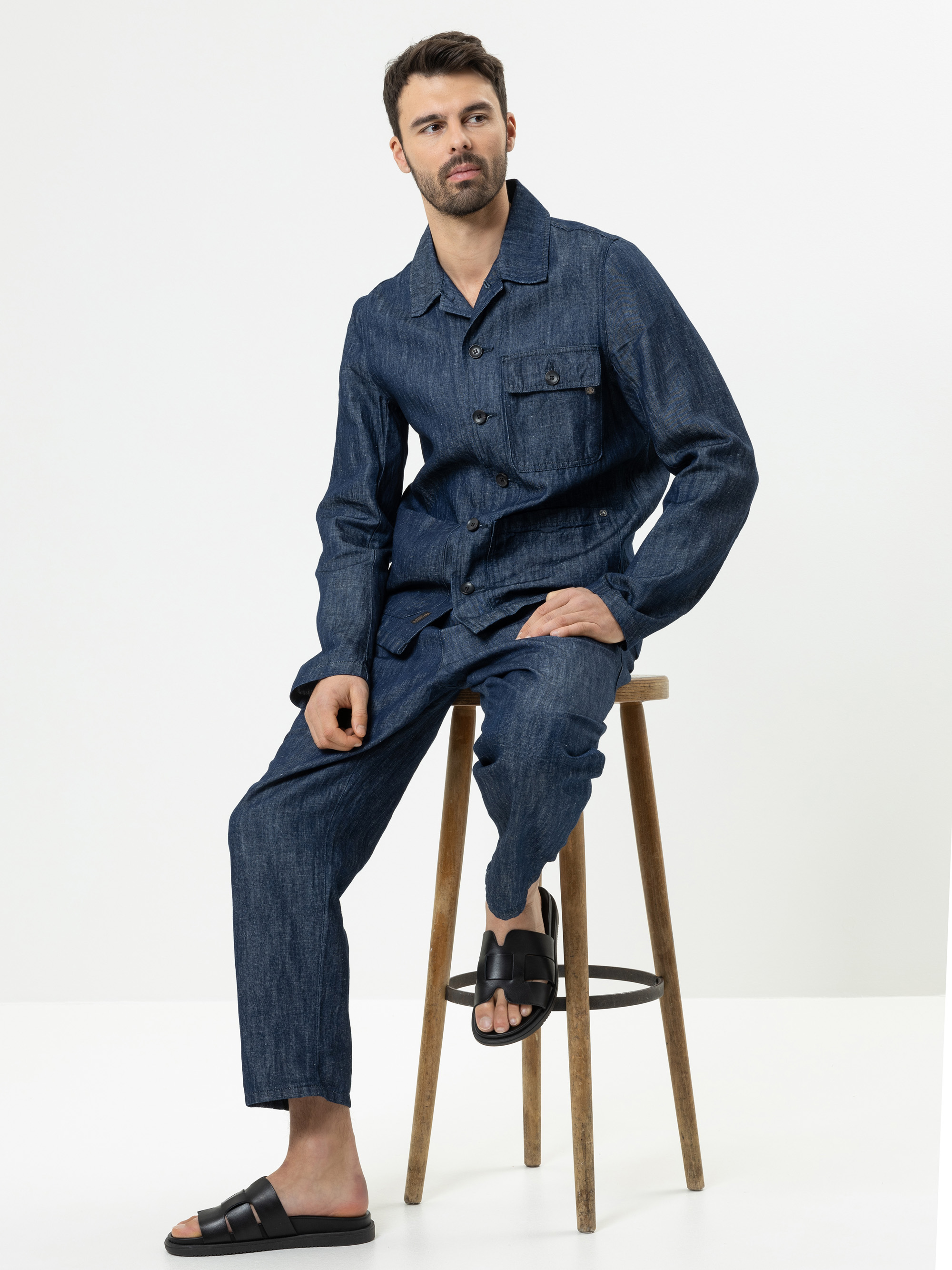 Ein Mann mit kurzen dunklen Haaren und einem Bart sitzt auf einem Holzschemel und trägt eine passende dunkle Jeansjacke und eine Hose mit schwarzen Slide-Sandalen. Der Hintergrund ist einfarbig weiß.