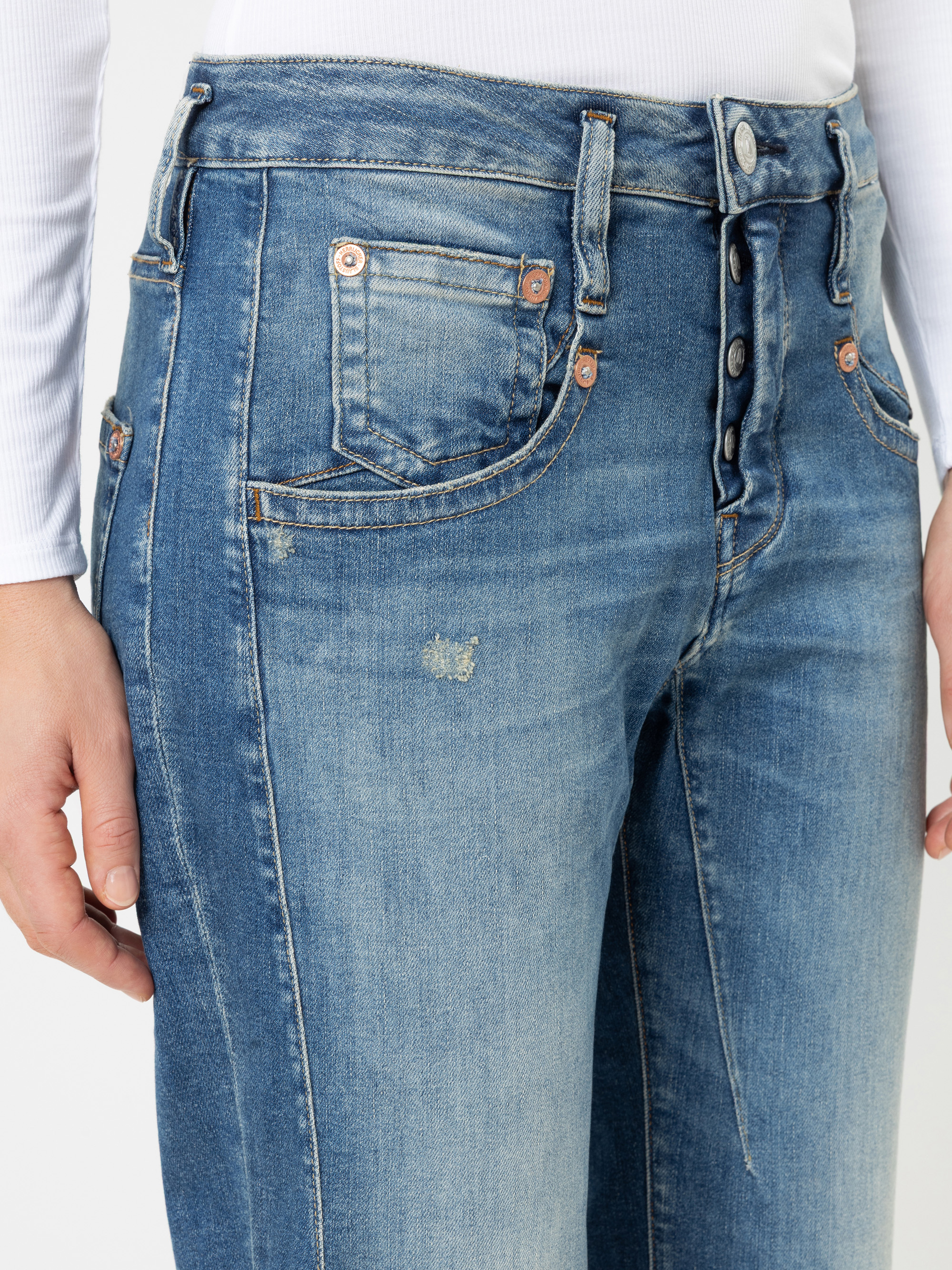 Nahaufnahme einer Person, die eine blaue Jeans mit hoher Taille und Knopfleiste sowie ein weißes Langarmhemd trägt. Die Jeans hat kupferfarbene Nieten und leichte Abnutzungsspuren im Bereich der Vordertasche.