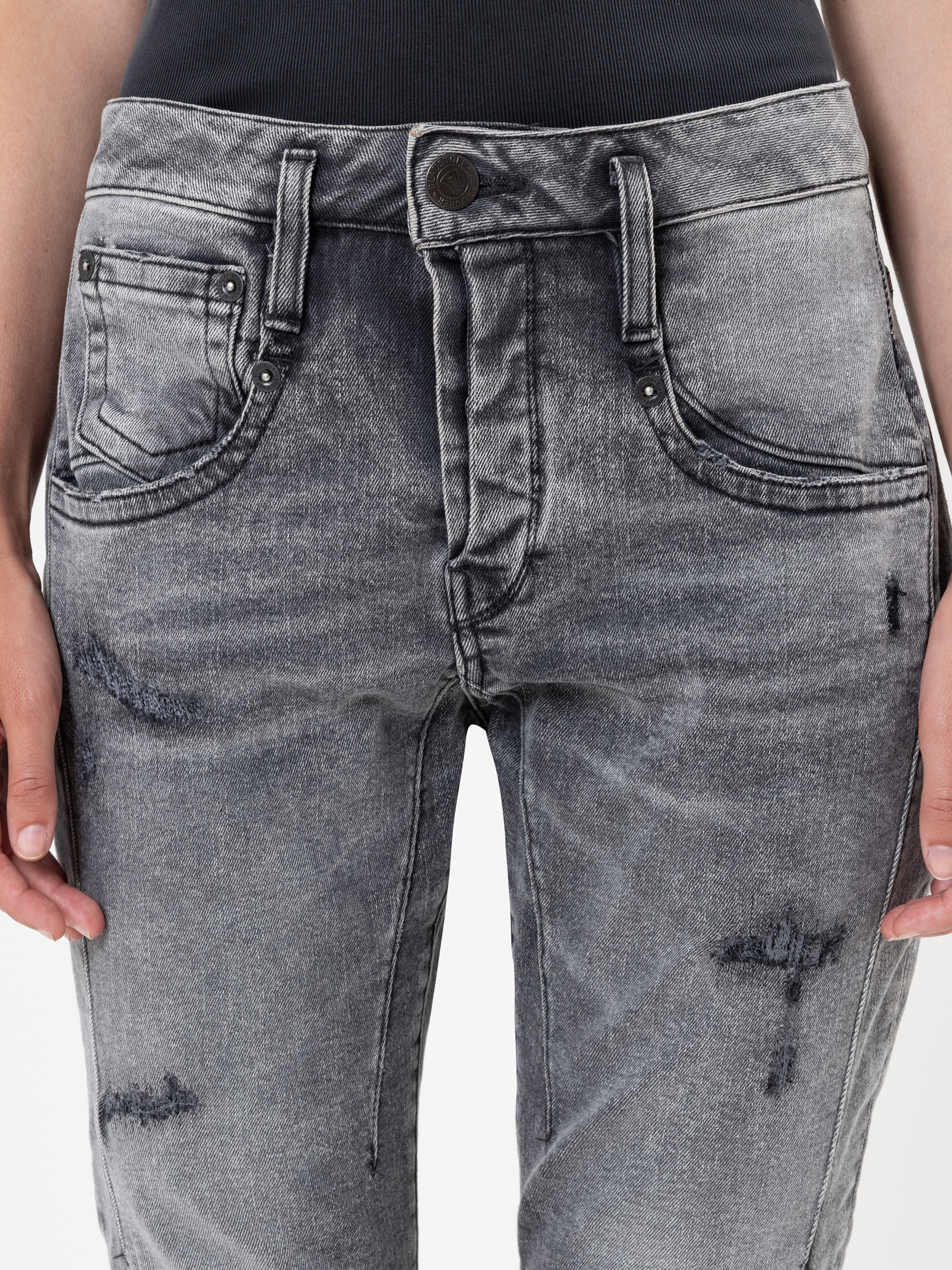 Nahaufnahme einer Person, die eine verblichene graue Jeans mit hoher Taille, schwarzem Knopf, Vordertaschen und gealterten Details trägt. Die Hände der Person liegen entspannt an ihren Seiten vor einem weißen Hintergrund.