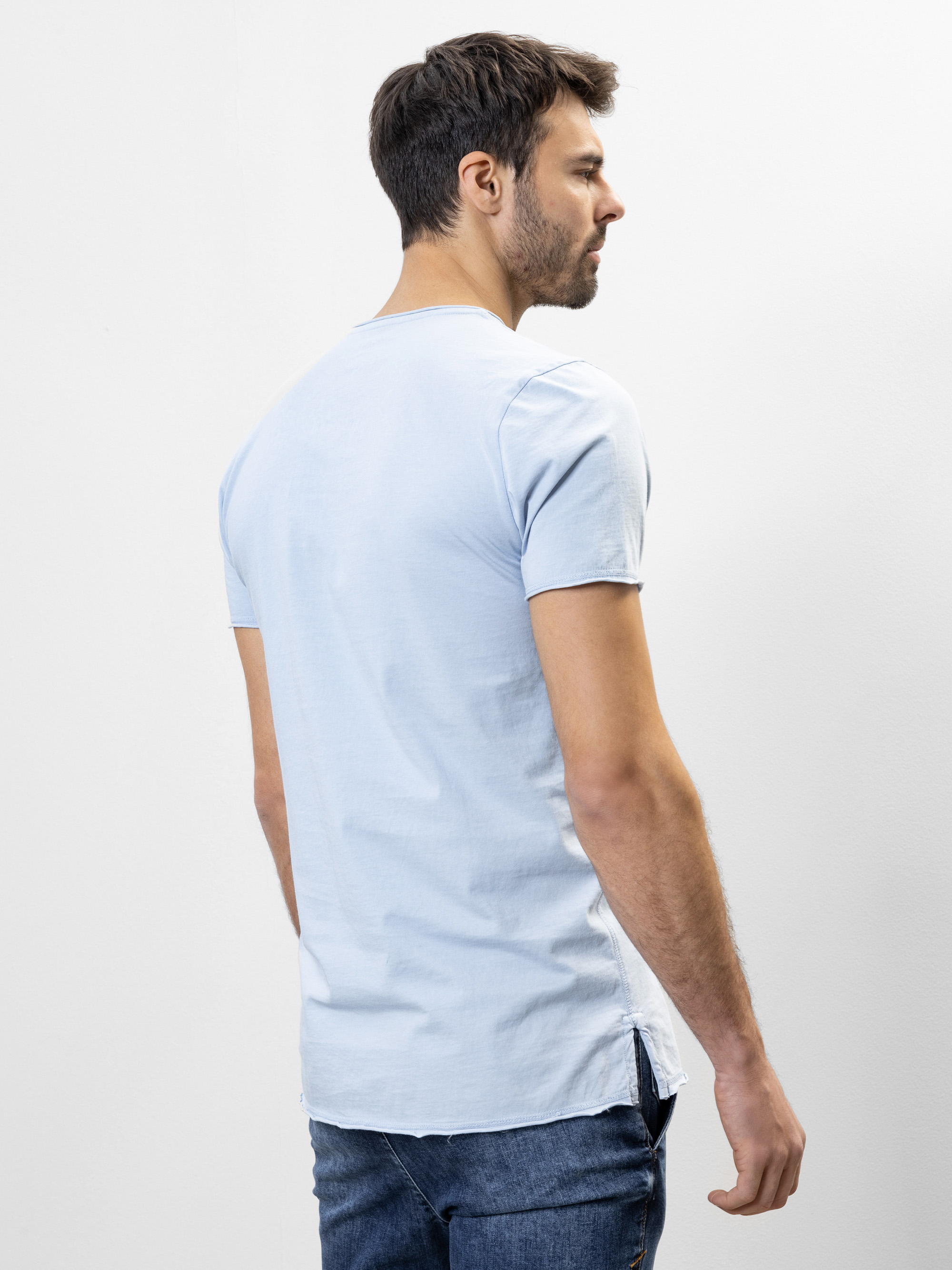 Ein Mann mit kurzen dunklen Haaren steht mit dem Gesicht zur Wand, trägt ein hellblaues, kurzärmeliges T-Shirt und blaue Jeans vor einem schlichten weißen Hintergrund.