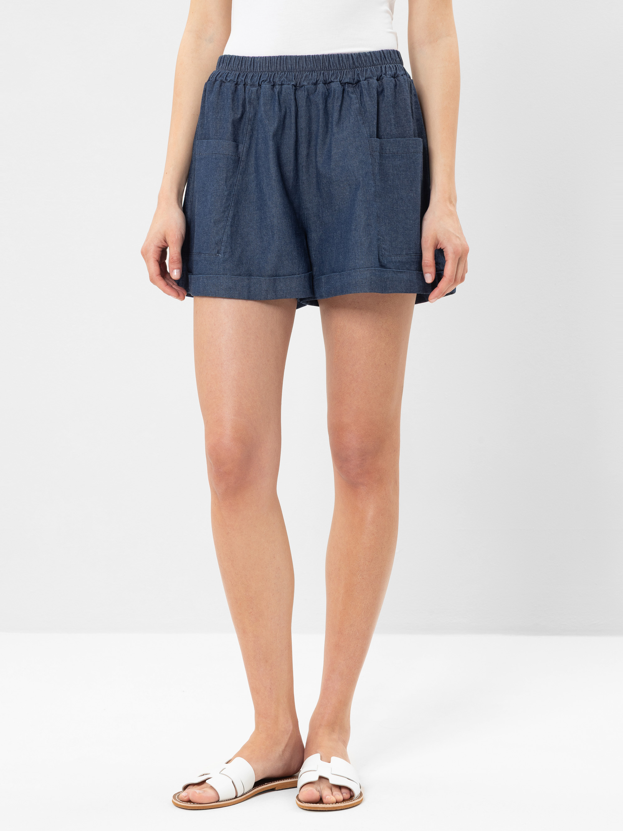 Eine Person, die ein weißes ärmelloses Oberteil, blaue Jeansshorts mit Vordertaschen und weiße Sandalen trägt, steht vor einem schlichten hellen Hintergrund.