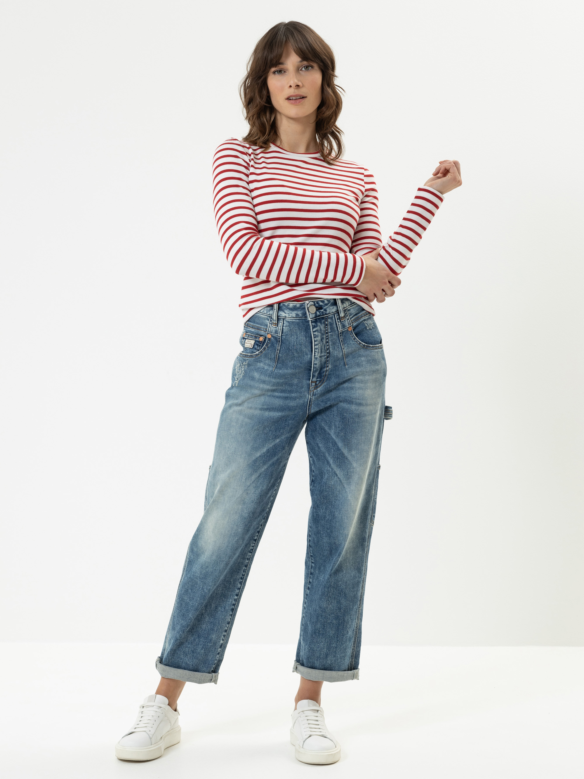 Eine Frau steht vor einem weißen Hintergrund und trägt ein rot-weiß gestreiftes, langärmeliges Hemd, lässige Blue Jeans und weiße Turnschuhe. Sie posiert mit einem angewinkelten Arm und einem lässigen Gesichtsausdruck.