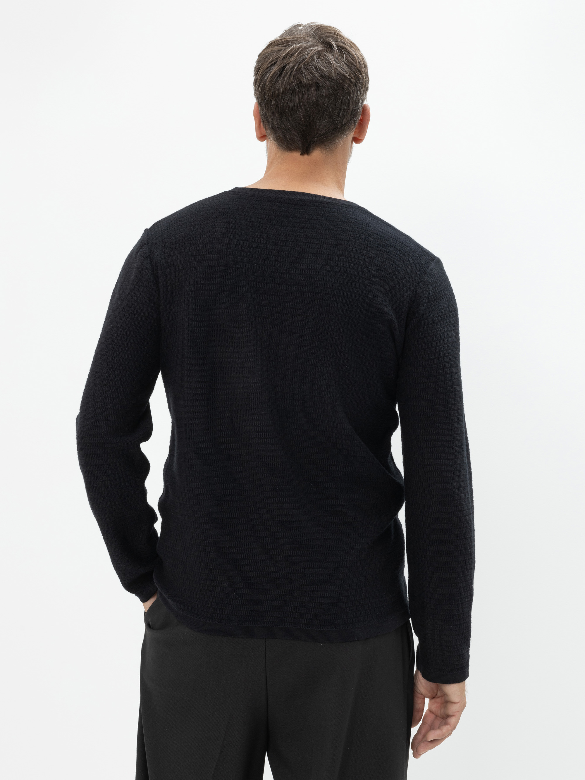 Ein Mann mit kurzen braunen Haaren steht mit dem Rücken zur Kamera, trägt einen langärmeligen schwarzen Pullover und eine schwarze Hose vor einem schlichten weißen Hintergrund.