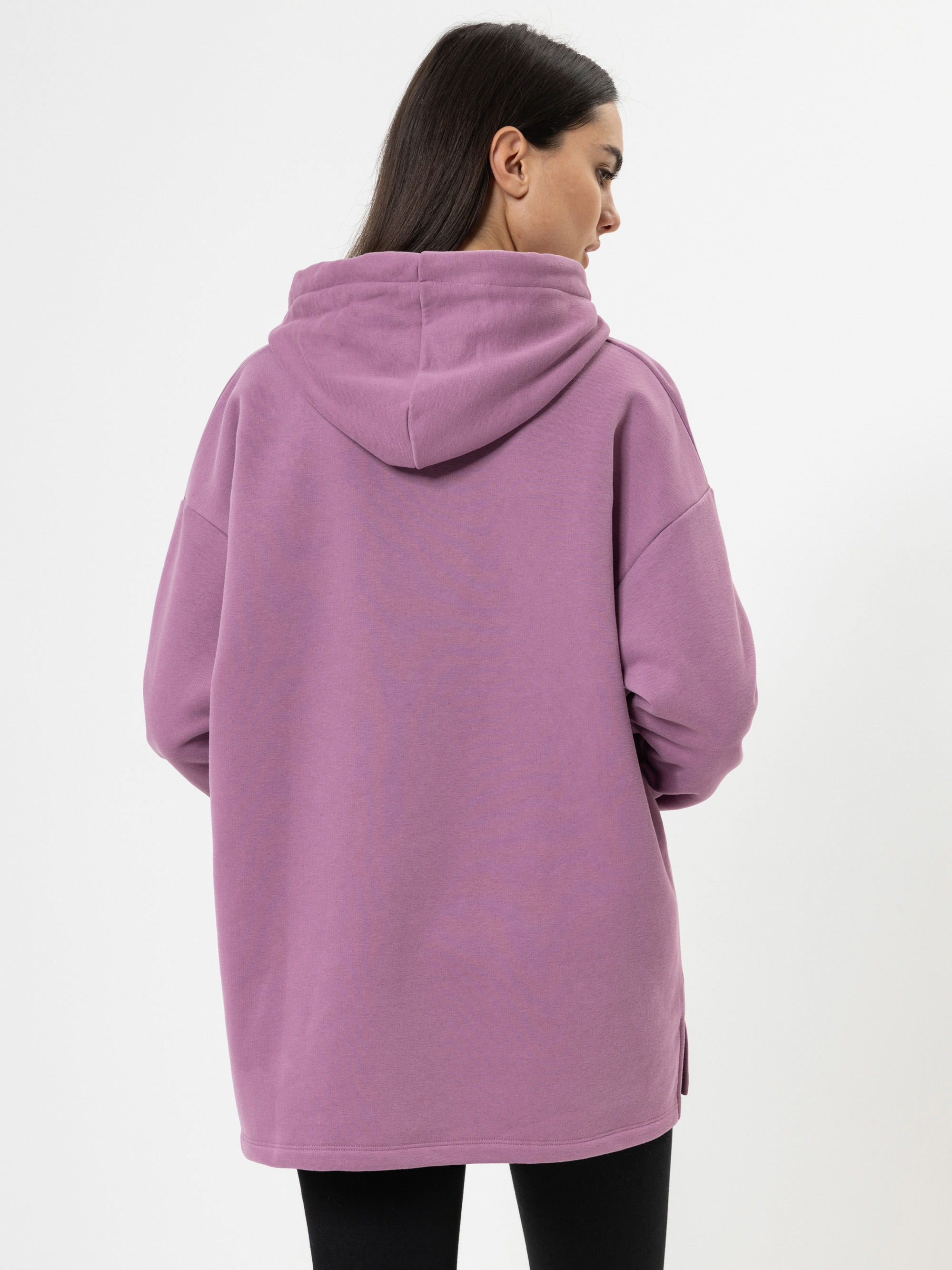 Eine Frau mit langen dunklen Haaren steht mit dem Rücken zur Kamera. Sie trägt einen übergroßen lila Kapuzenpulli und eine schwarze Hose vor einem schlichten weißen Hintergrund.