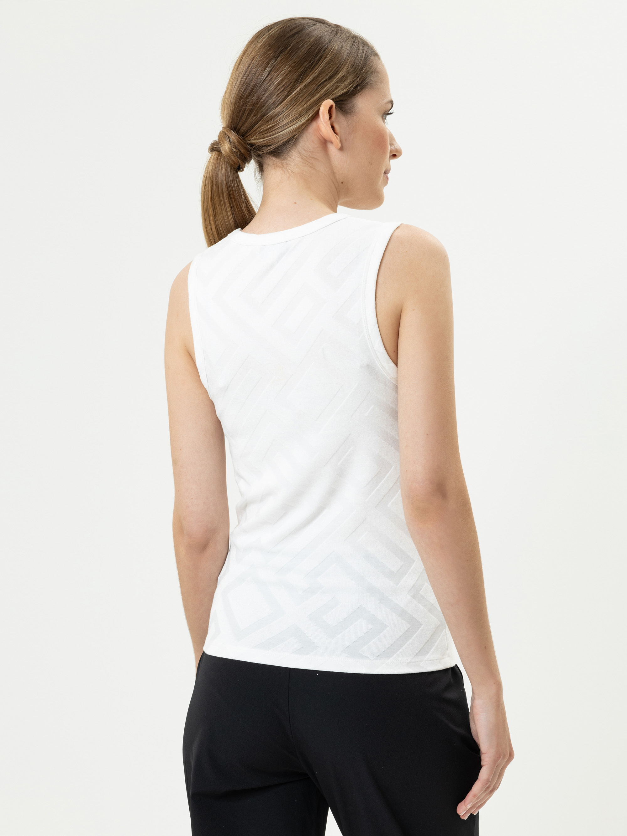 Eine Frau mit hellbraunem Haar in einem niedrigen Pferdeschwanz trägt ein ärmelloses weißes Oberteil mit einem dezenten geometrischen Muster und eine schwarze Hose, die von der Kamera weg vor einem weißen Hintergrund steht.